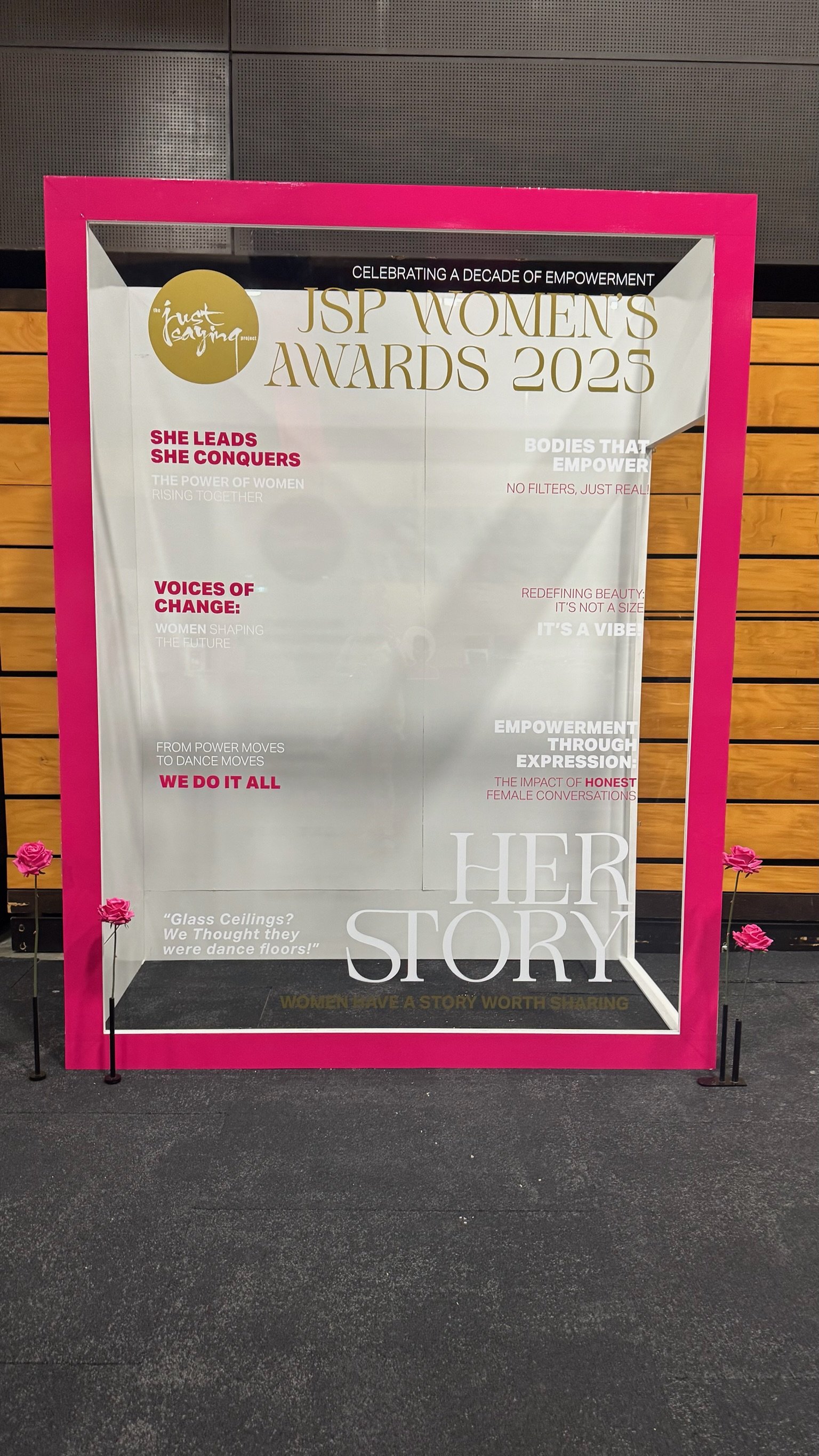 A pink-framed glass display with text and pink roses on a black surface. The display features information about the JSP Women's Awards 2025, floral decorations, and inspirational quotes celebrating women's stories and empowerment.