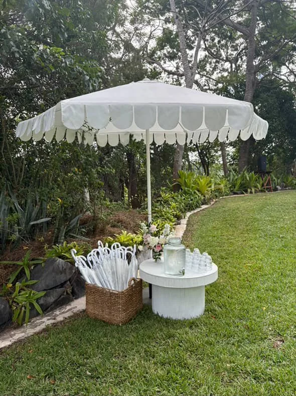 Outdoor setup with a white round table, a large white umbrella, a basket of white plastic utensils, a glass jar with flowers, and a row of small glasses on the table. Green grass and trees in the background.