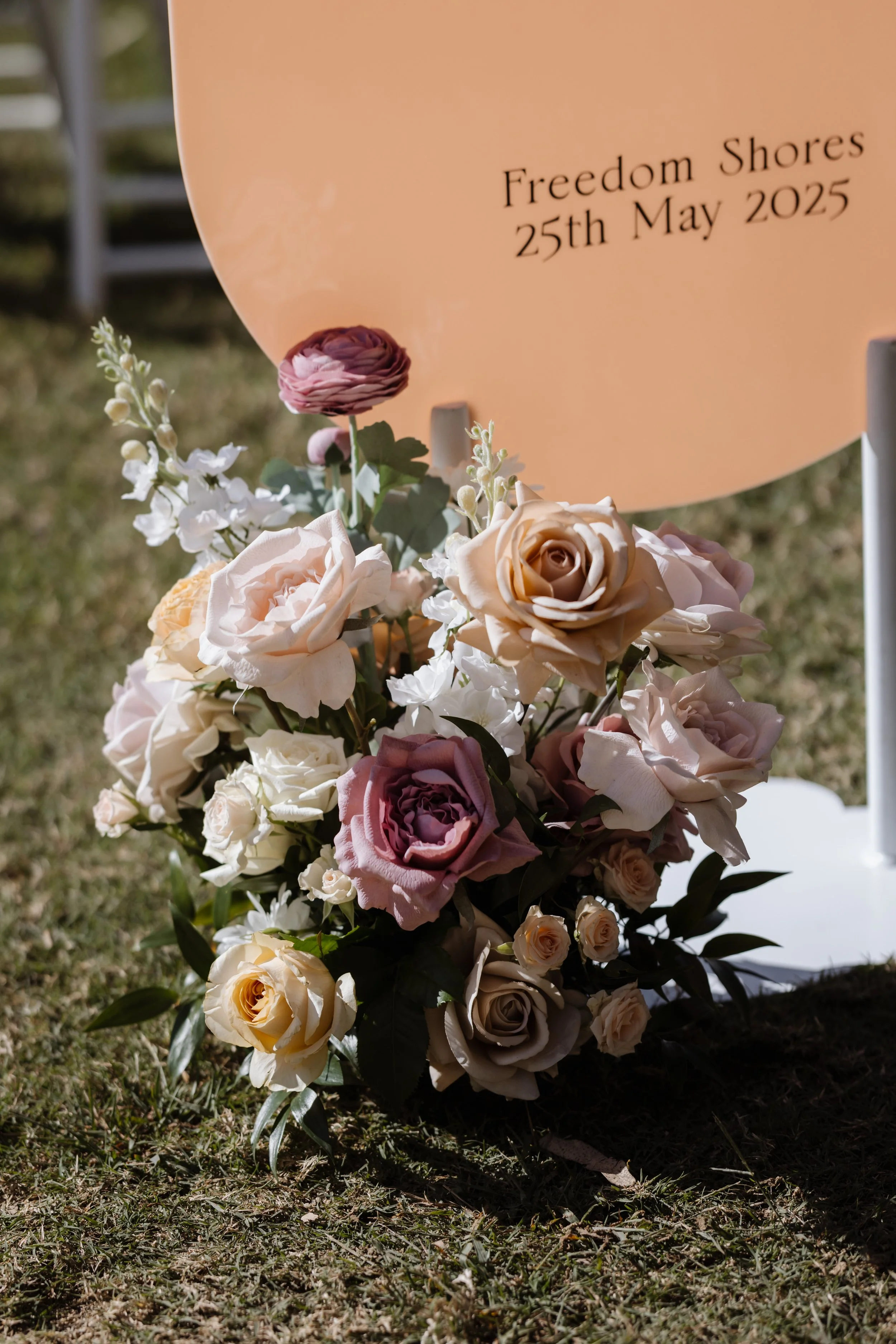 A floral arrangement with roses and other flowers in pastel colors placed on grass, with a memorial sign reading 'Freedom Shores 25th May 2025' in the background.