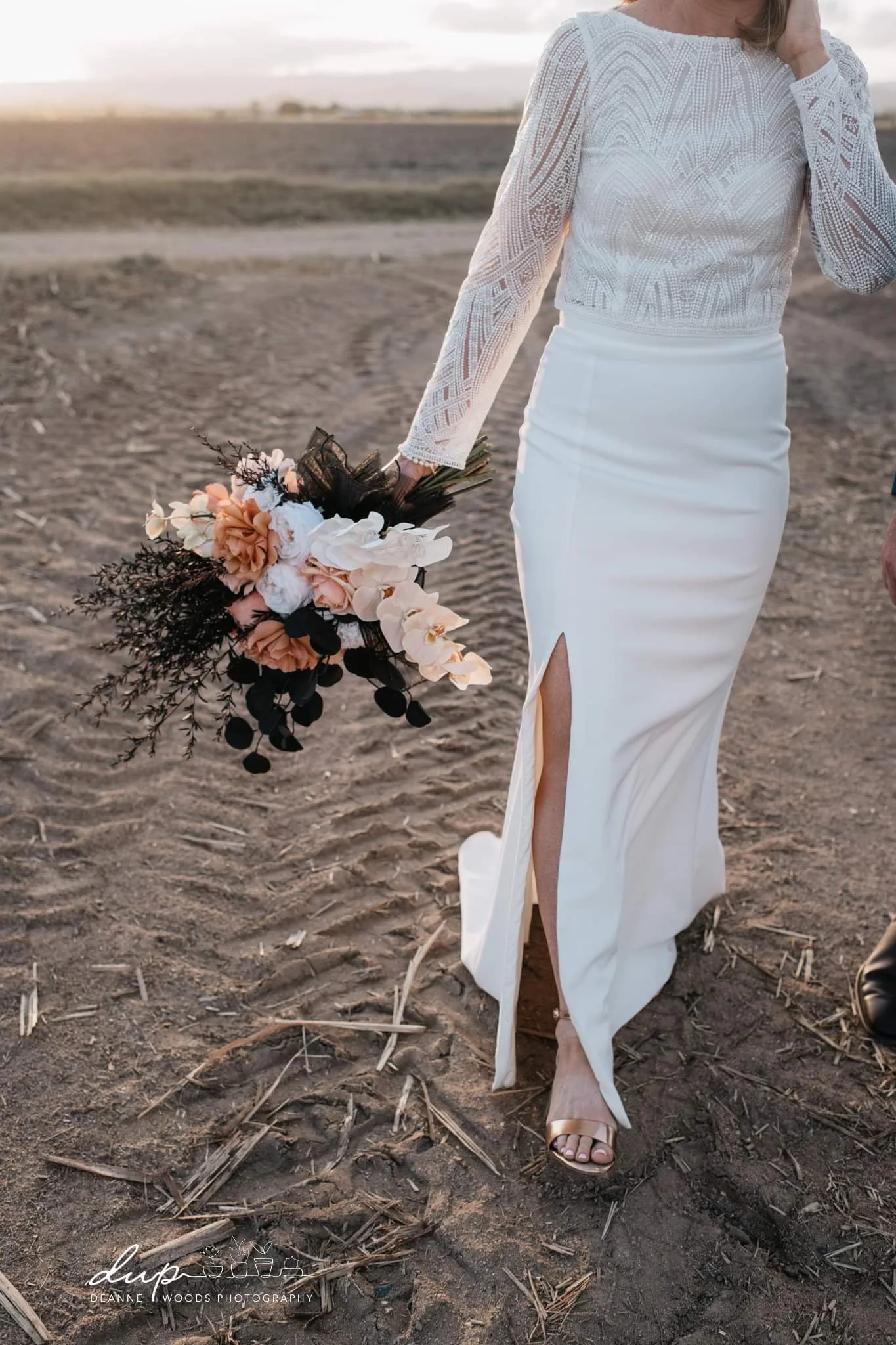 Close-up of a woman in a white wedding dress holding a bouquet of flowers. She is walking on a dirt path outdoors at sunset, wearing open-toe heels with a slit in her dress revealing her leg.