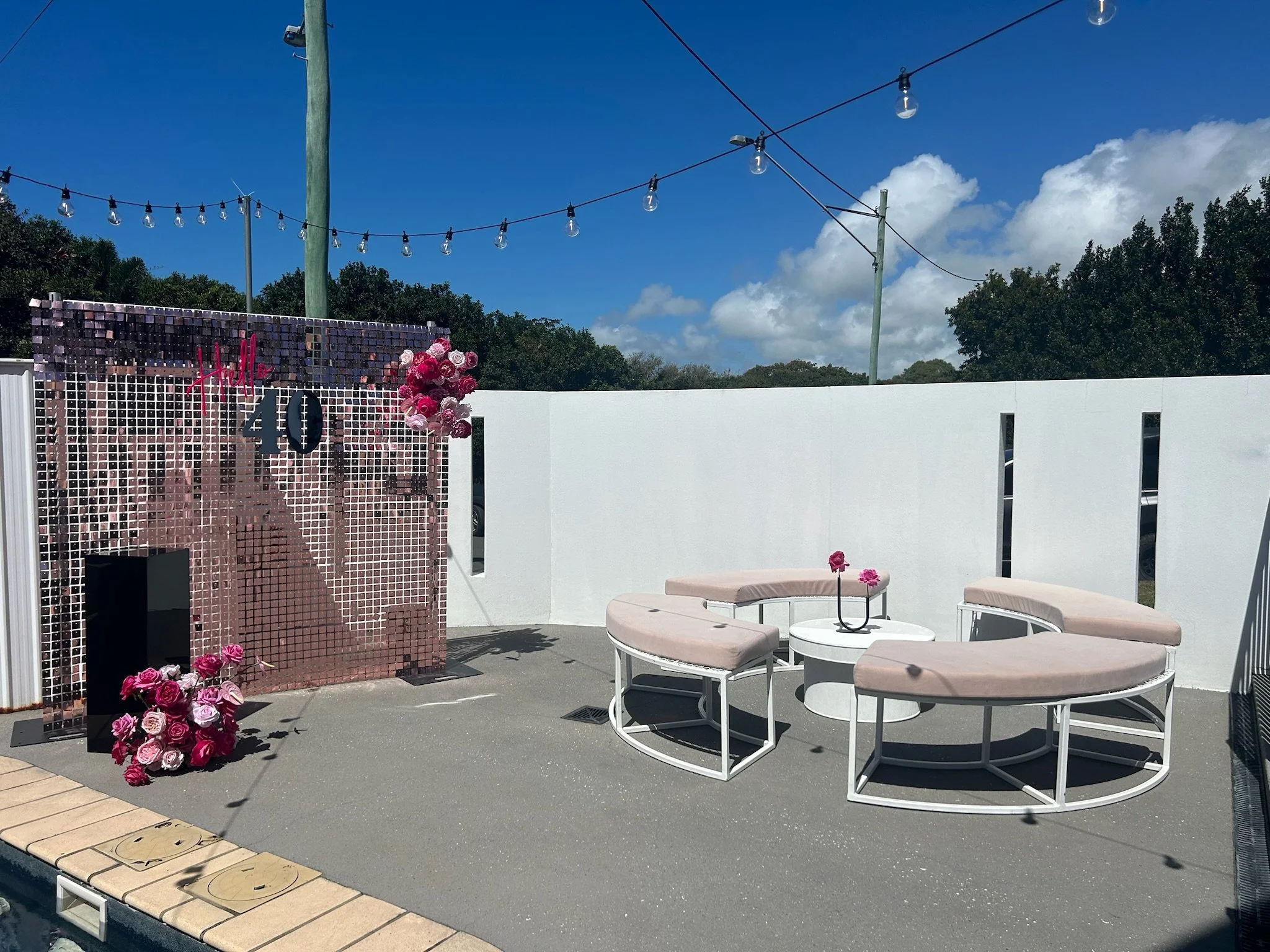 Outdoor patio with pink seating area, white table, decorative pink flowers, and a shimmering pink mosaic wall with 