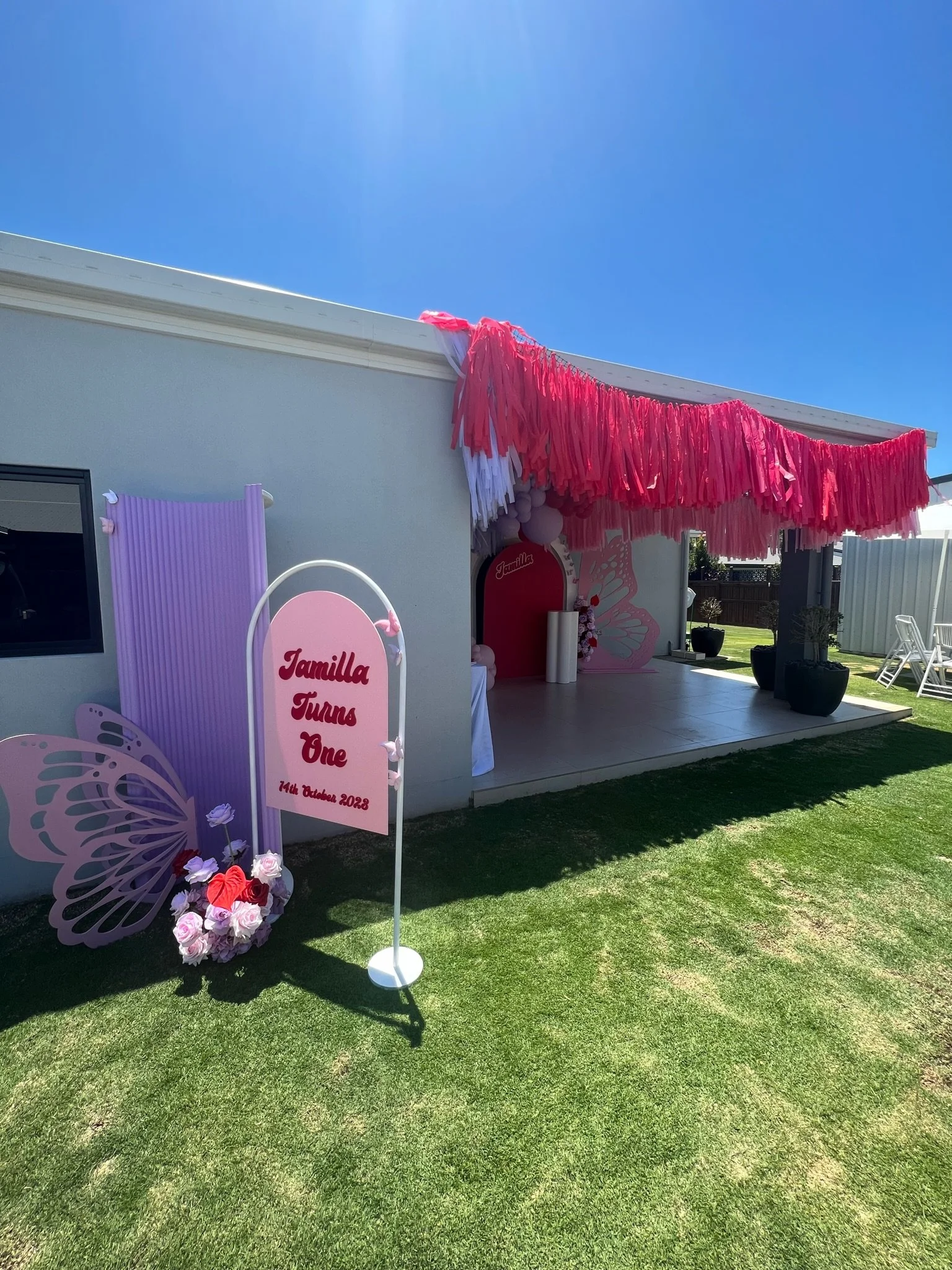 Decorated outdoor area for a first birthday celebration with pink and purple butterfly theme, a sign reading 'Jamilla Turns One, 14th October 2023,' pink and white paper tassels, and flower arrangements.