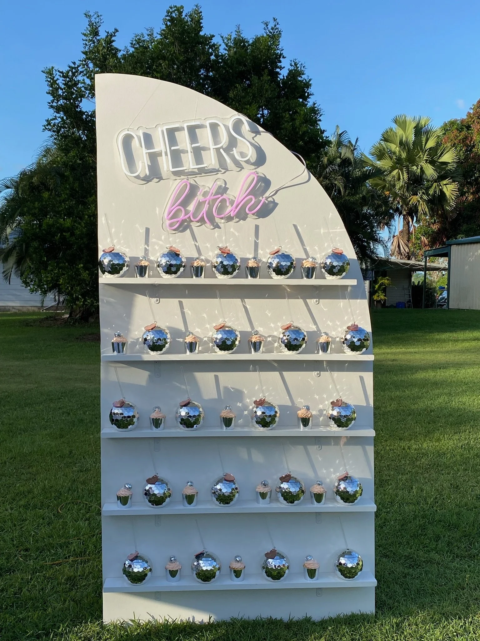 A decorative display with a sign that says 'CHEERS b*tch' in neon lights, surrounded by shiny, reflective ornaments and small cupcake decorations, set outdoors on green grass with trees in the background.