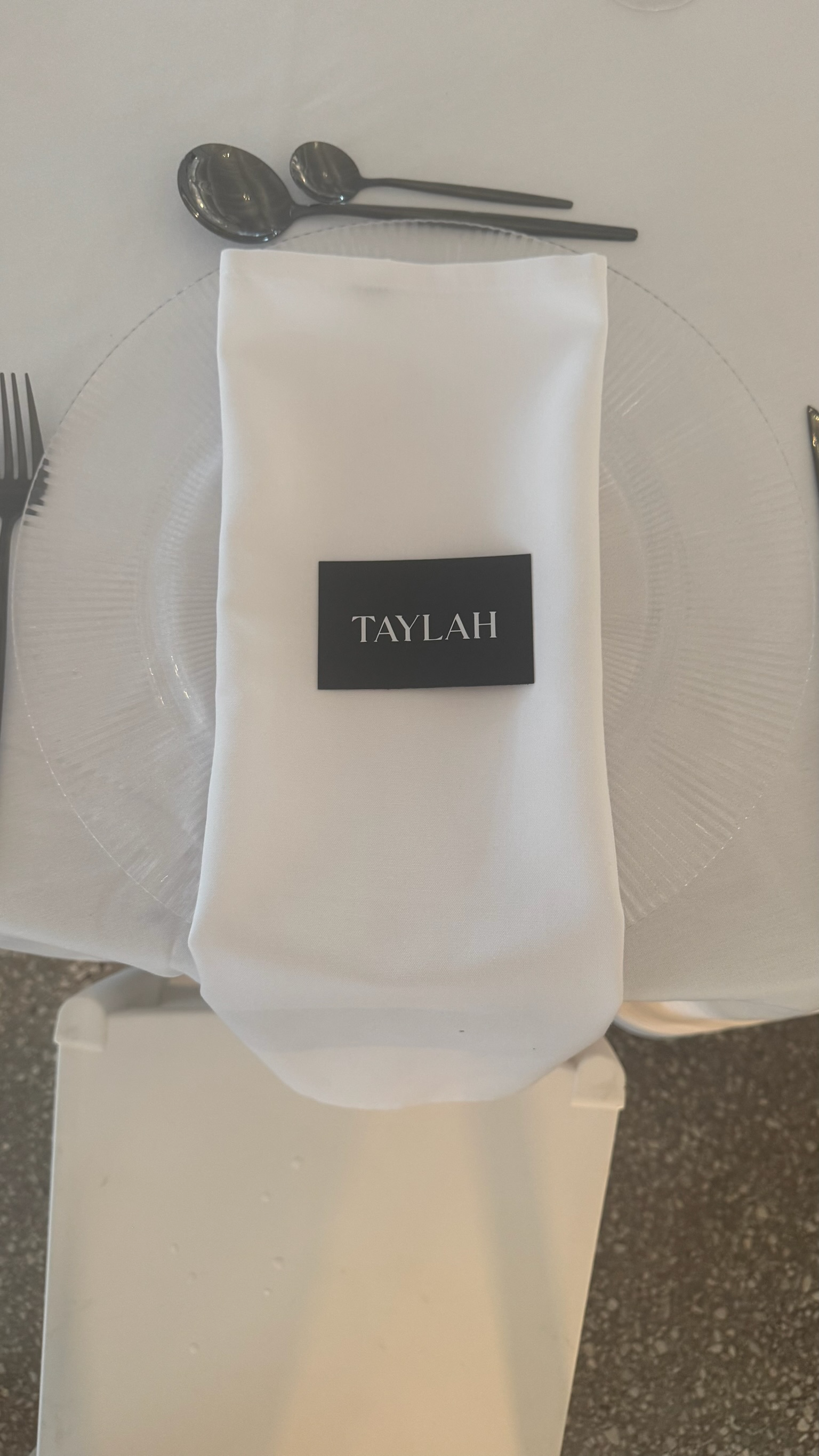 Place setting with a white napkin, black place card with the name 'TAYLAH', clear glass plate, and silverware on a white tablecloth.