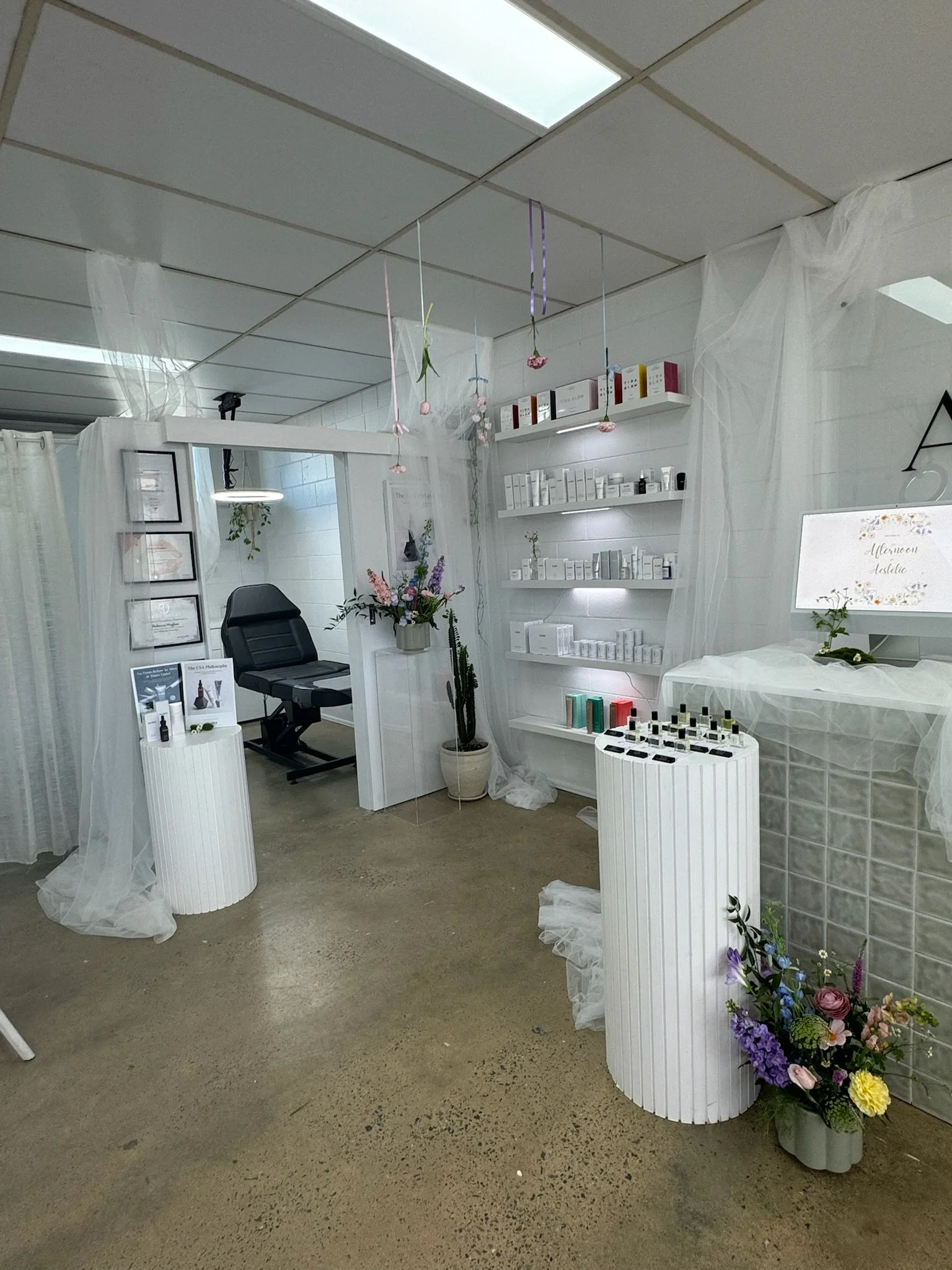 Interior of a spa or beauty clinic with a white and minimalist decor, shelves filled with skincare products, a reception desk with flowers, and a treatment room with a reclining chair, decorated with sheer fabric and hanging decorations.