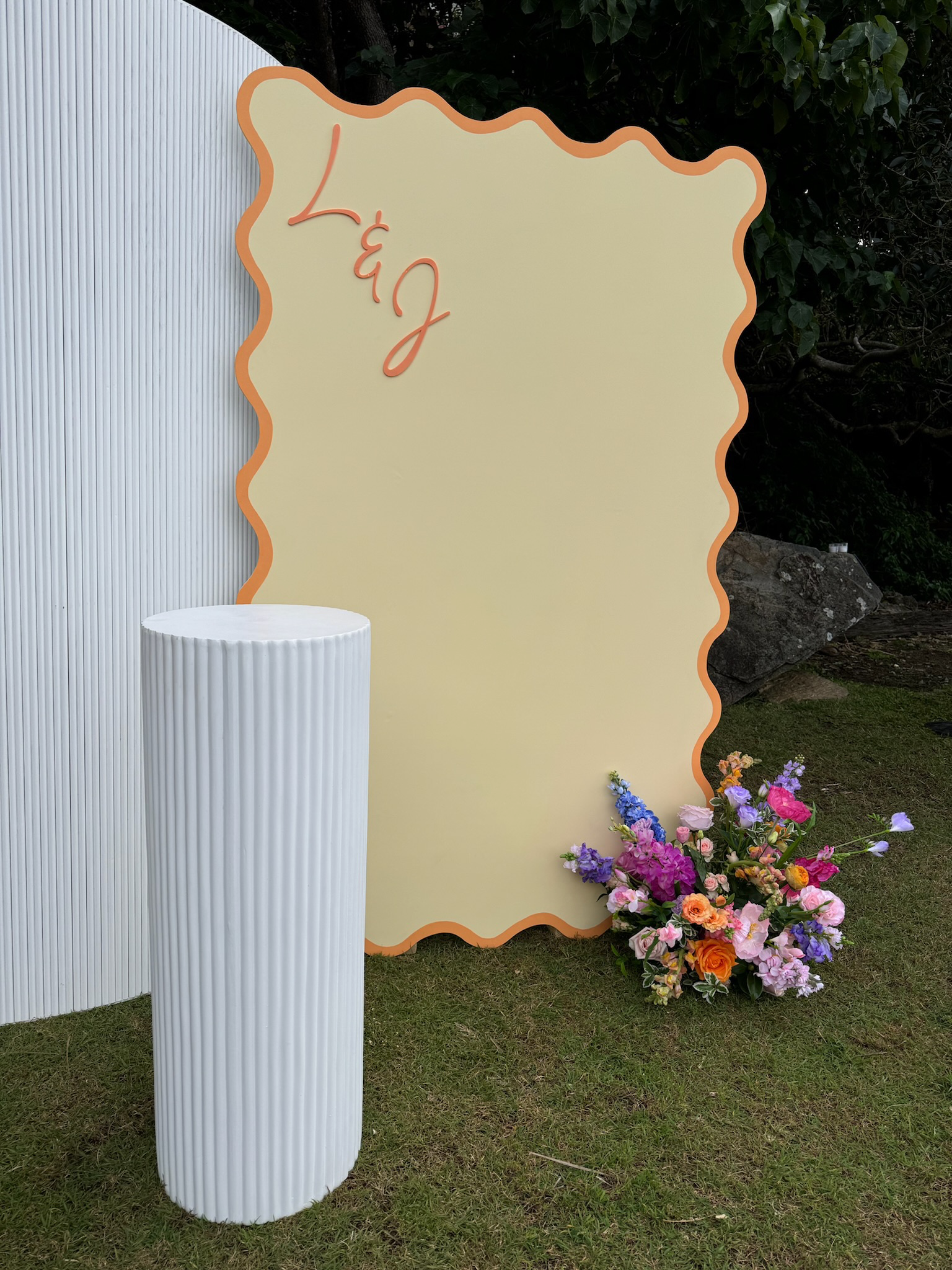 A yellow decorative board with orange wavy border and the word 'Love' written in cursive, placed outdoors near a flower arrangement with pink, purple, and orange flowers, and a white ribbed cylindrical stand on grass.
