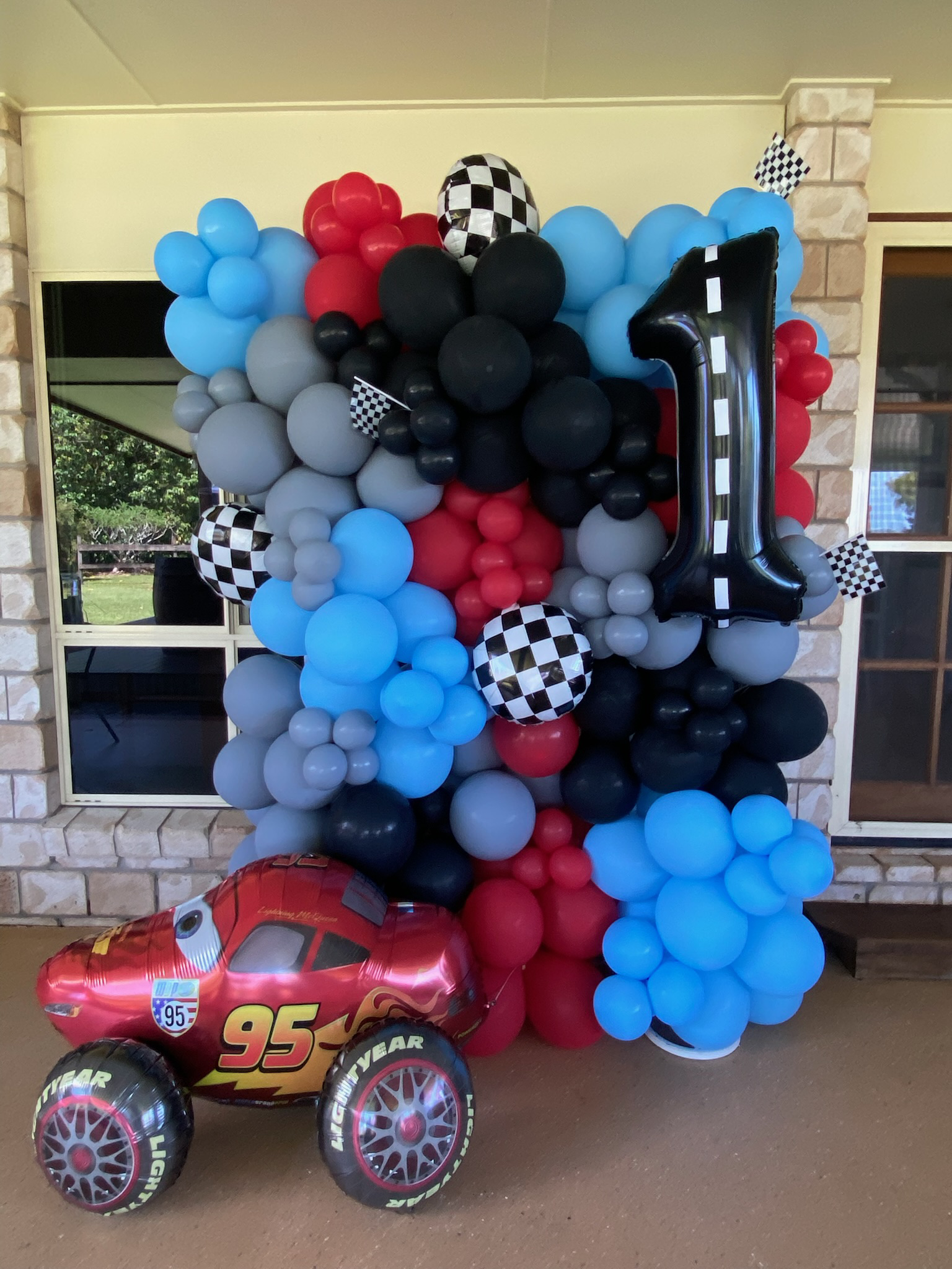 A colorful balloon arrangement featuring red, black, gray, and blue balloons with checkered flags, a large number one balloon, and a Lightning McQueen car-shaped balloon. The display appears to be for a first birthday celebration.