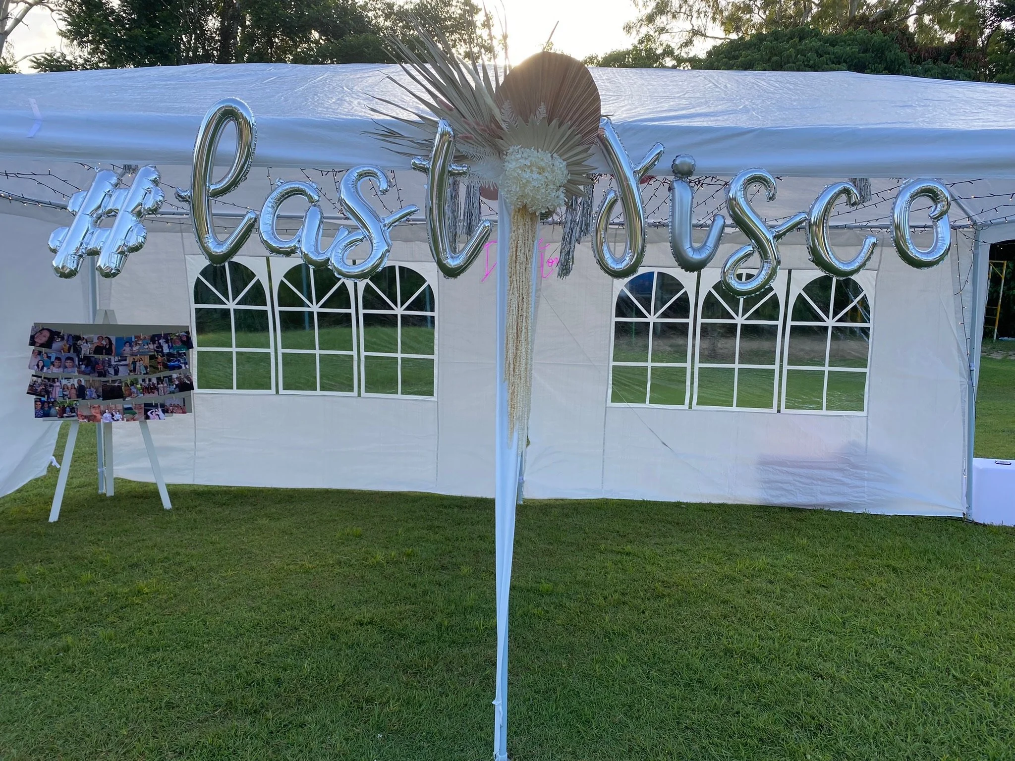 White event tent with silver balloon letters spelling #LasDucos, decorated with flowers and greenery, with a photo collage on A-frame easel, and green grass on ground.