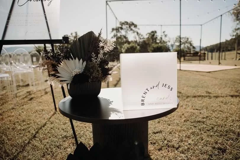 A round black table with a floral arrangement in a dark vase and a white card with handwritten names, set outdoors on a grassy field with a sports net and bleachers in the background.