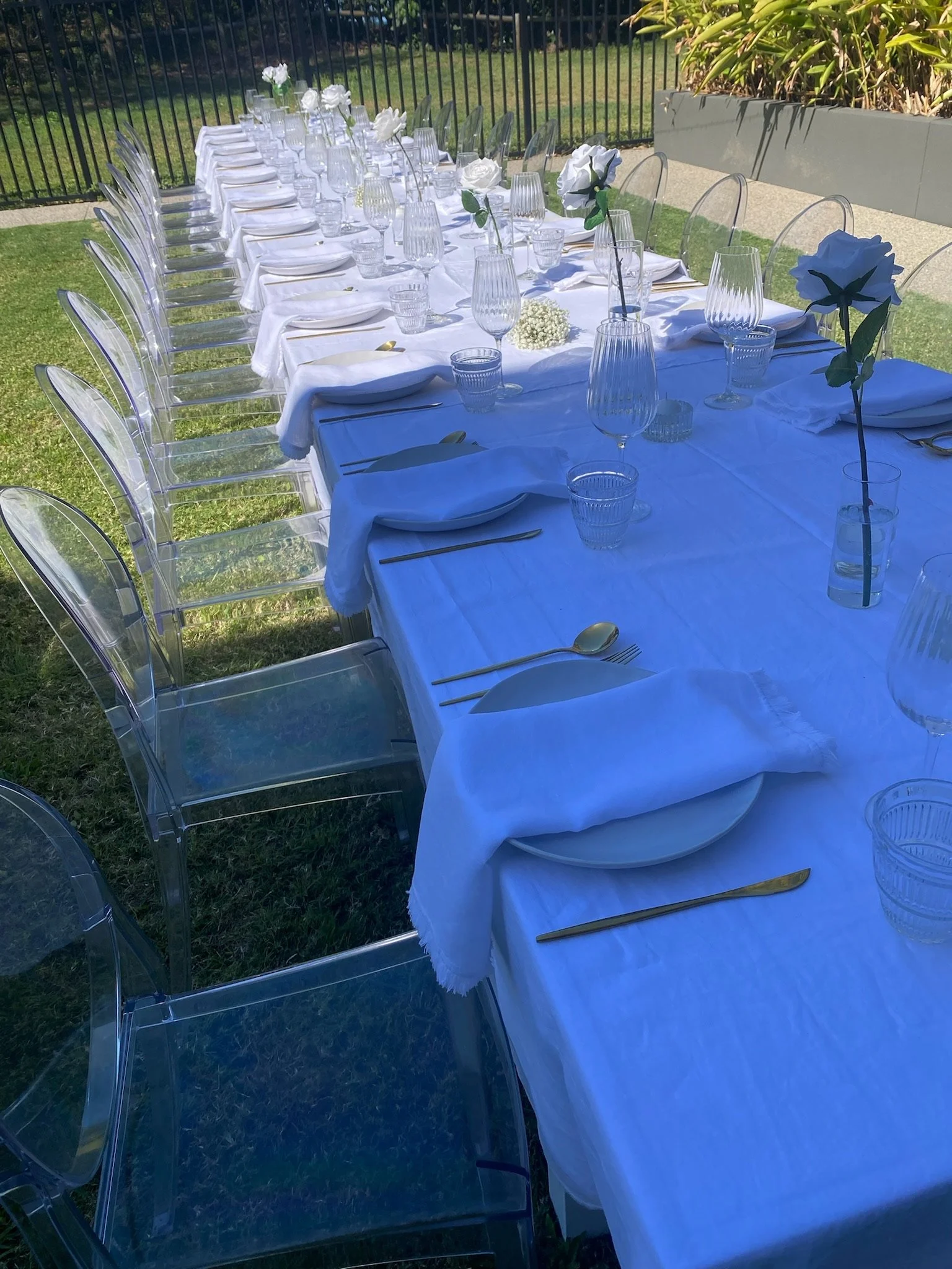Outdoor dining table set with white tablecloth, white napkins, glassware, and gold cutlery, surrounded by transparent chairs, with floral centerpieces and a fenced garden background.
