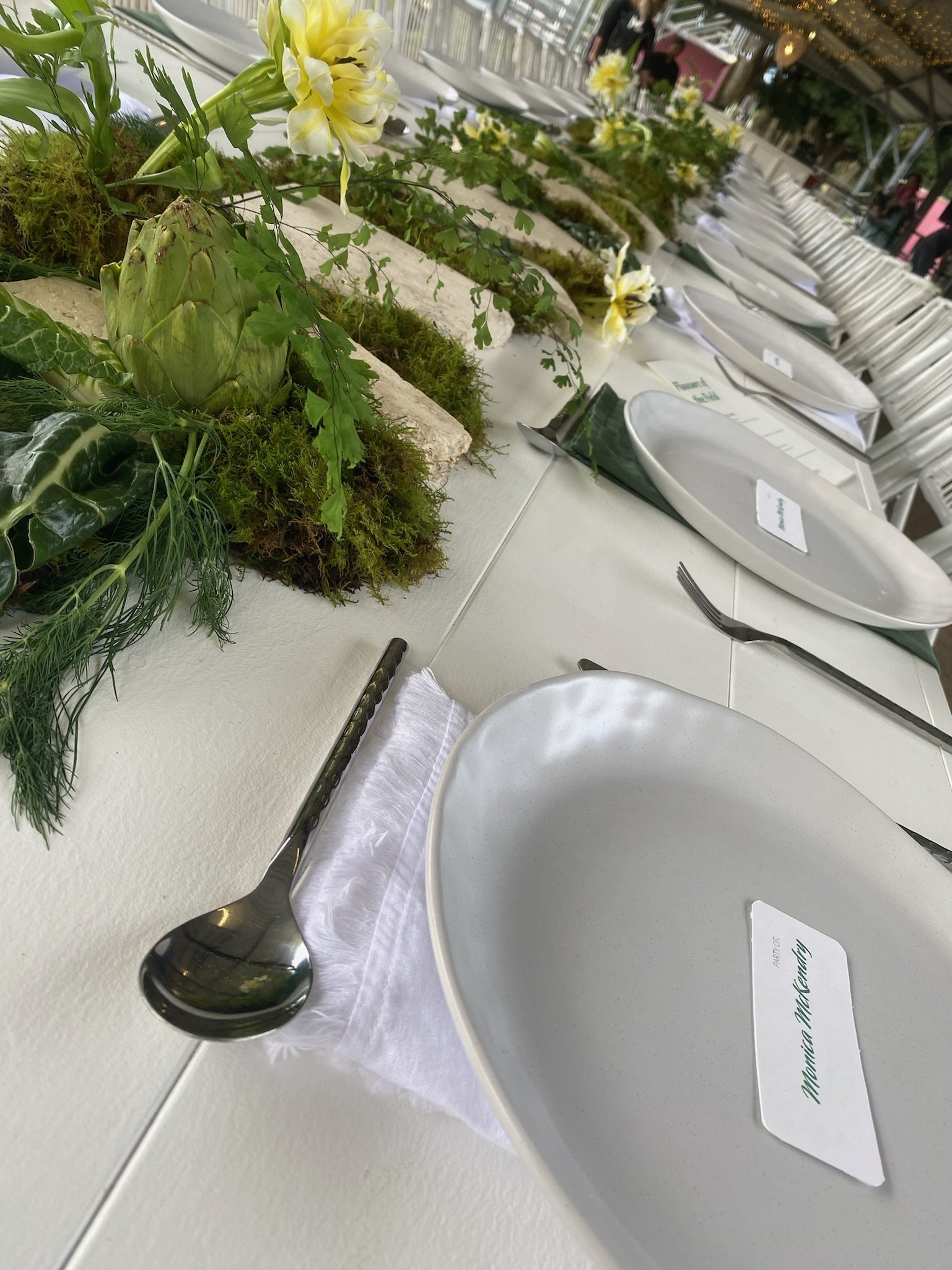 Empty white dinner plate and silver spoon on a white napkin at a decorated table with greenery and yellow flowers.