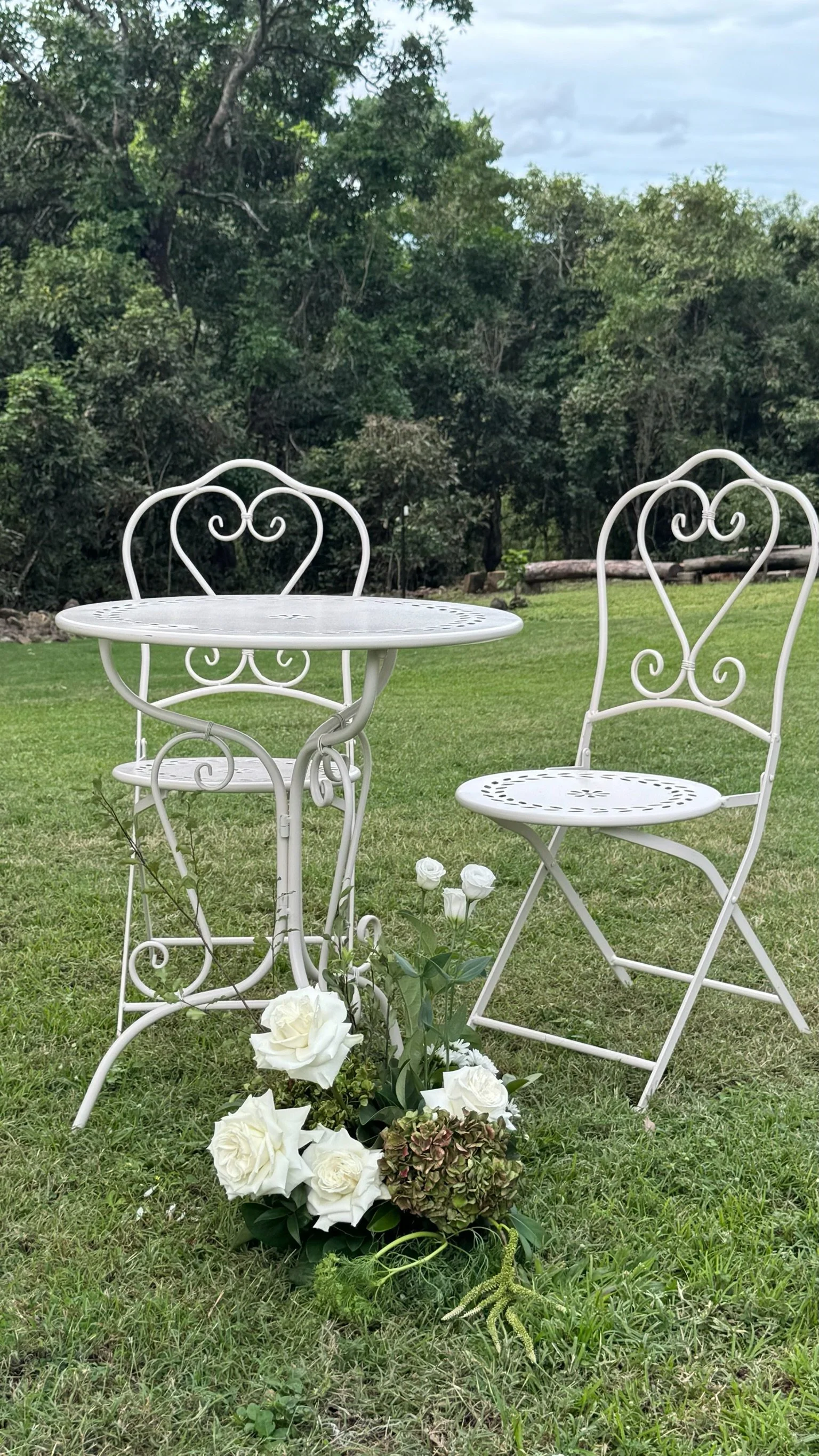 White wrought iron outdoor table and chair set with a flower arrangement on the grass in a lush green garden.