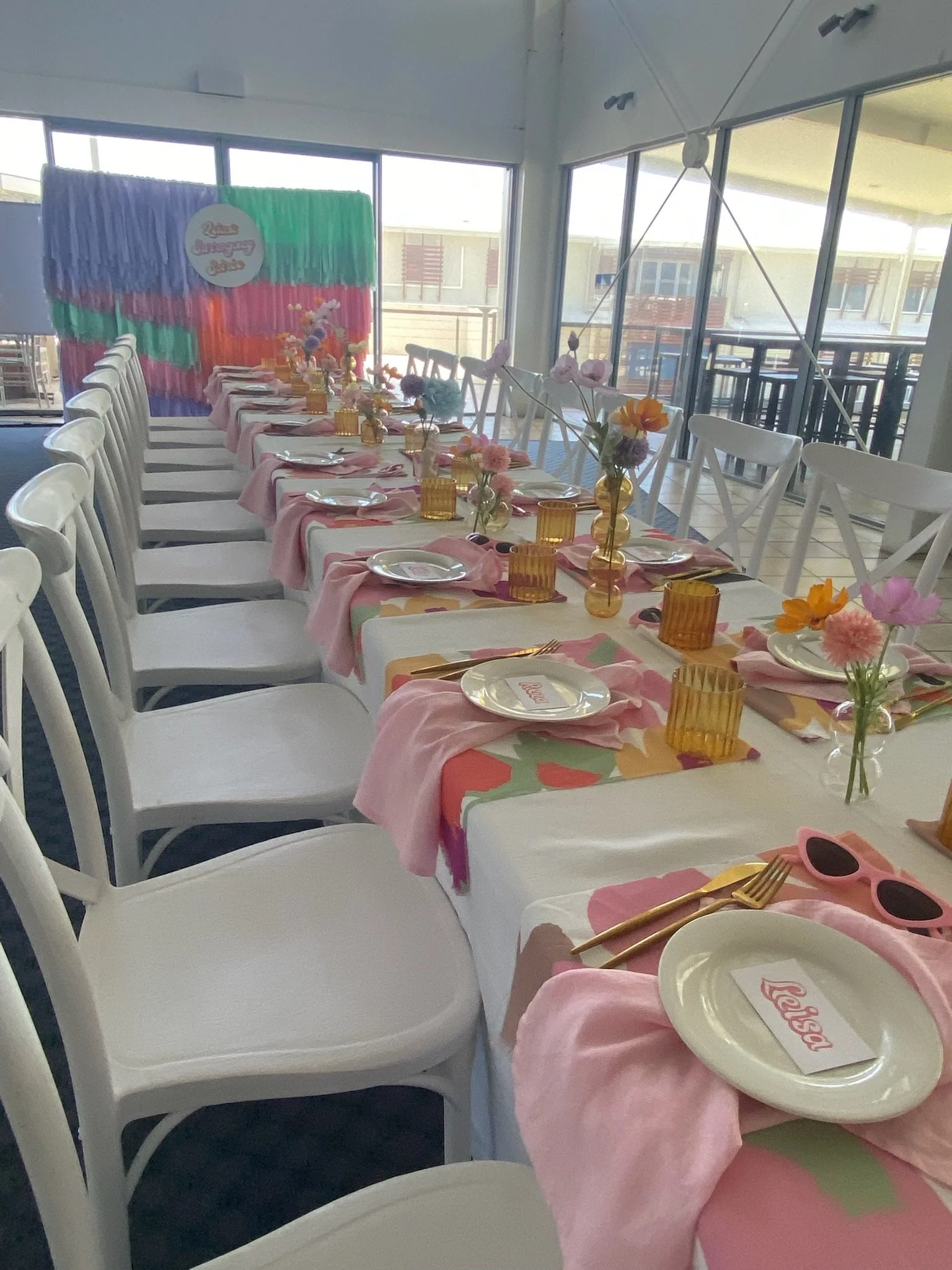 A long table set for a celebration with pink napkins, floral centerpieces, yellow glasses, white plates with pink 'Baby' cards, and gold utensils. The background features a colorful backdrop with a label that reads 'Baby Shower' in pink and purple.