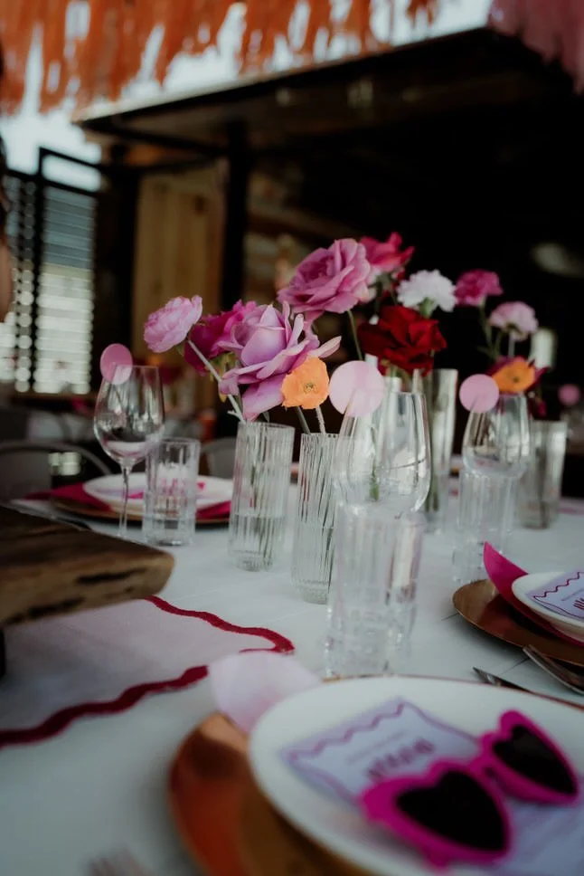 Table decor with pink, purple, and red flowers in glass vases at a dining table with wine glasses, plates, and pink sunglasses.