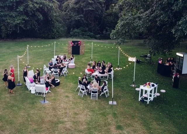 Outdoor party with groups of people sitting at white tables under string lights, on a grassy field with trees in the background, and a photo booth and a DJ setup nearby.