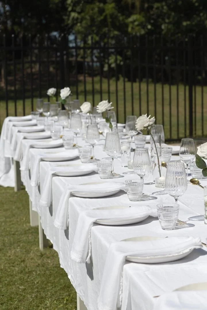 A long outdoor banquet table set with white tablecloths, plates, glasses, and white flowers in vases, with a grassy area and fence in the background.