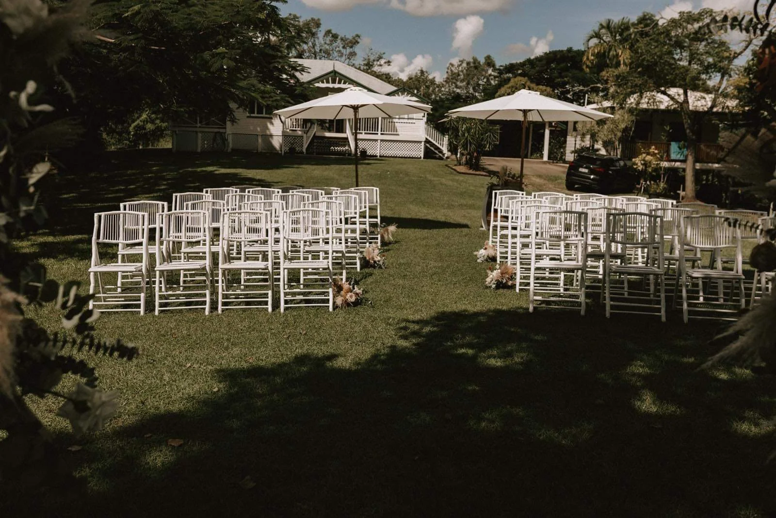 Outdoor wedding setup with rows of white chairs and floral arrangements, shaded by large umbrellas on a grassy lawn.
