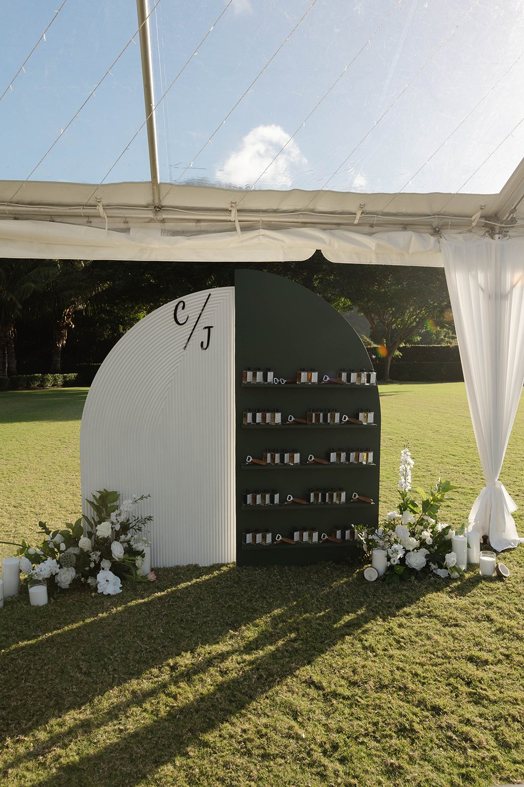 Outdoor wedding or event decor with a black and white display shelf under a white tent, surrounded by floral arrangements and candles, on a sunny day in a grassy area with trees in the background.