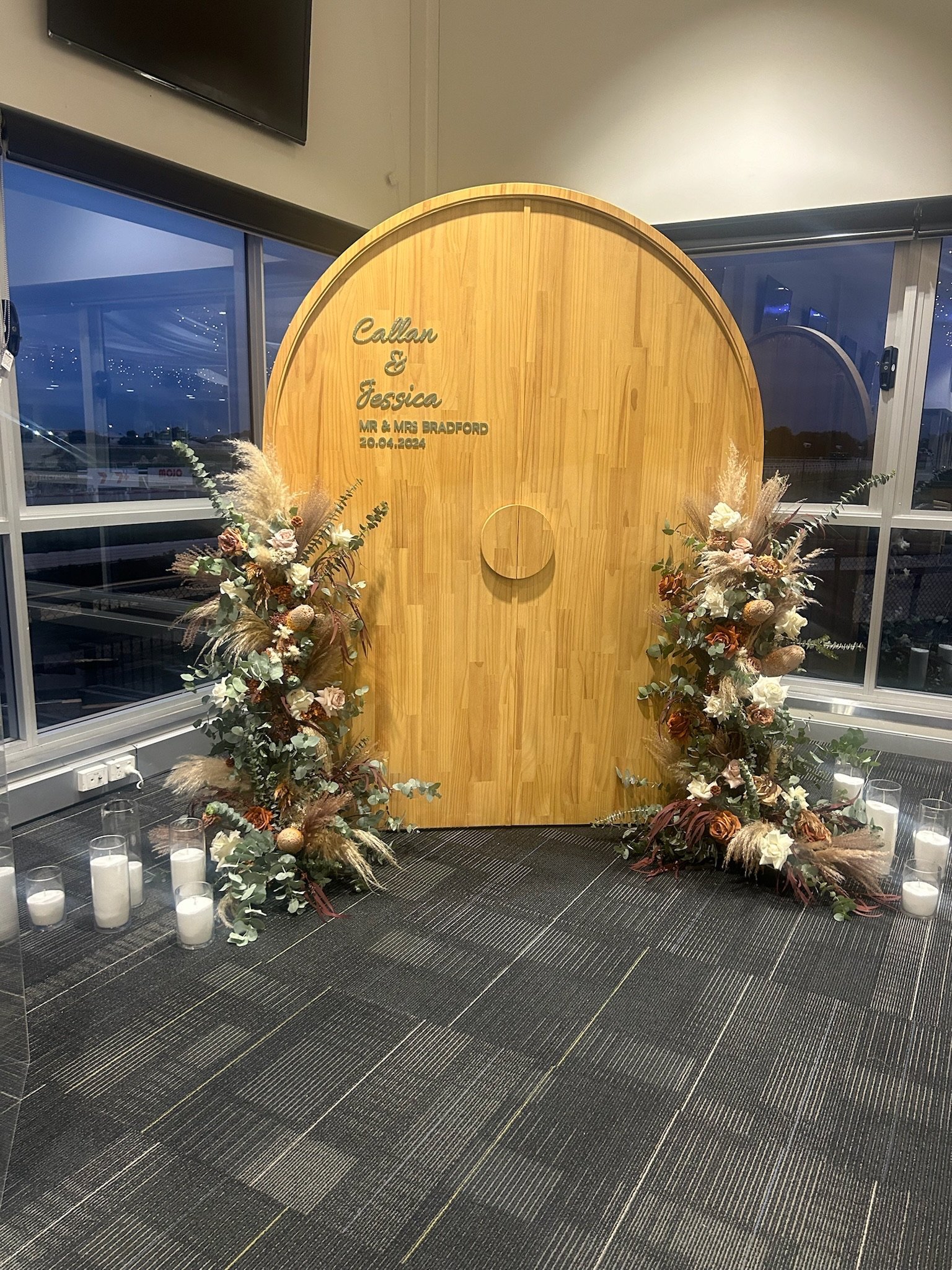 Decorative wooden panel with engraved text, flanked by two floral arrangements with candles on the floor.
