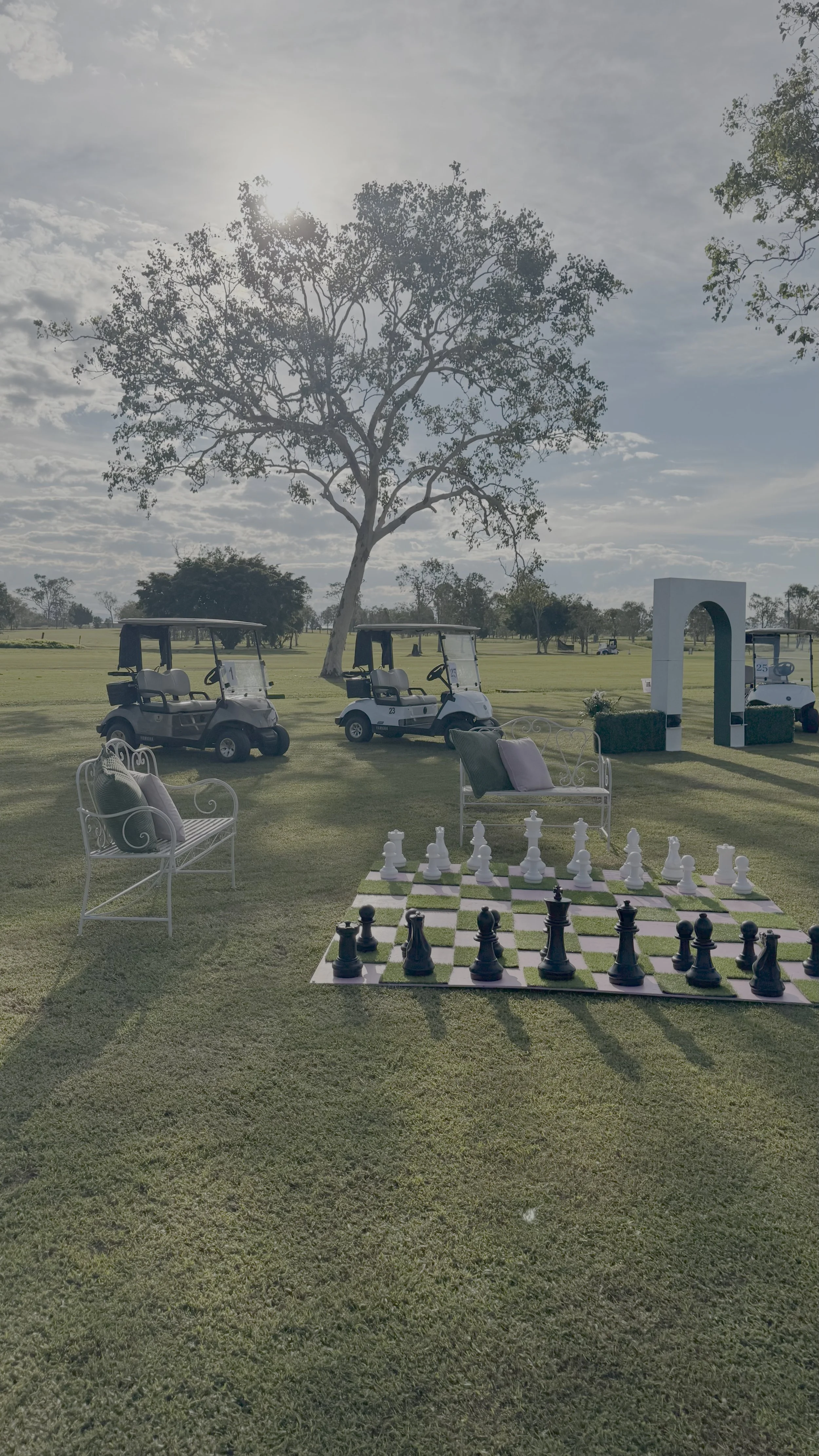 A golf course with a large tree in the background, golf carts parked, and a chessboard with large chess pieces set up on the grass in the foreground. White benches with pillows sit nearby, along with a decorative archway.