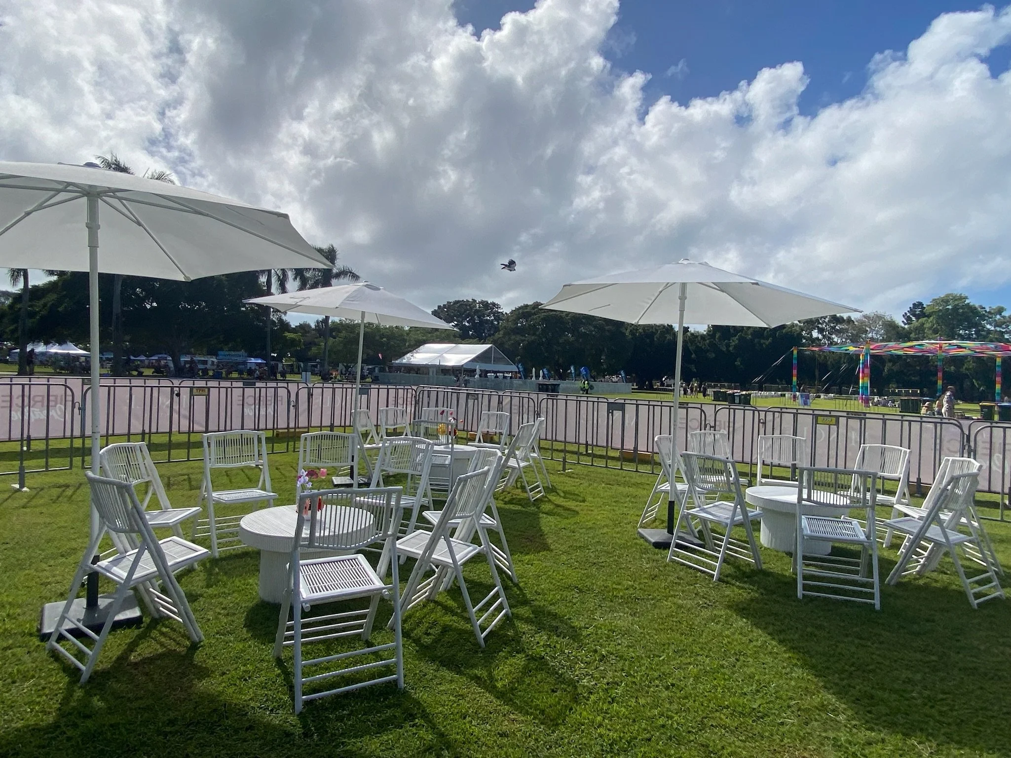 Outdoor event space with white chairs and tables under large white umbrellas on a grassy area, surrounded by metal barricades, with tents, colorful rides, and a partially cloudy sky in the background.