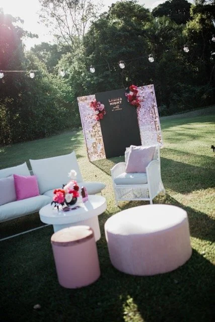 Outdoor event setup with white furniture, pink cushions, flower arrangements, a backdrop with 'Lush Night' written on it, string lights, and trees in the background.