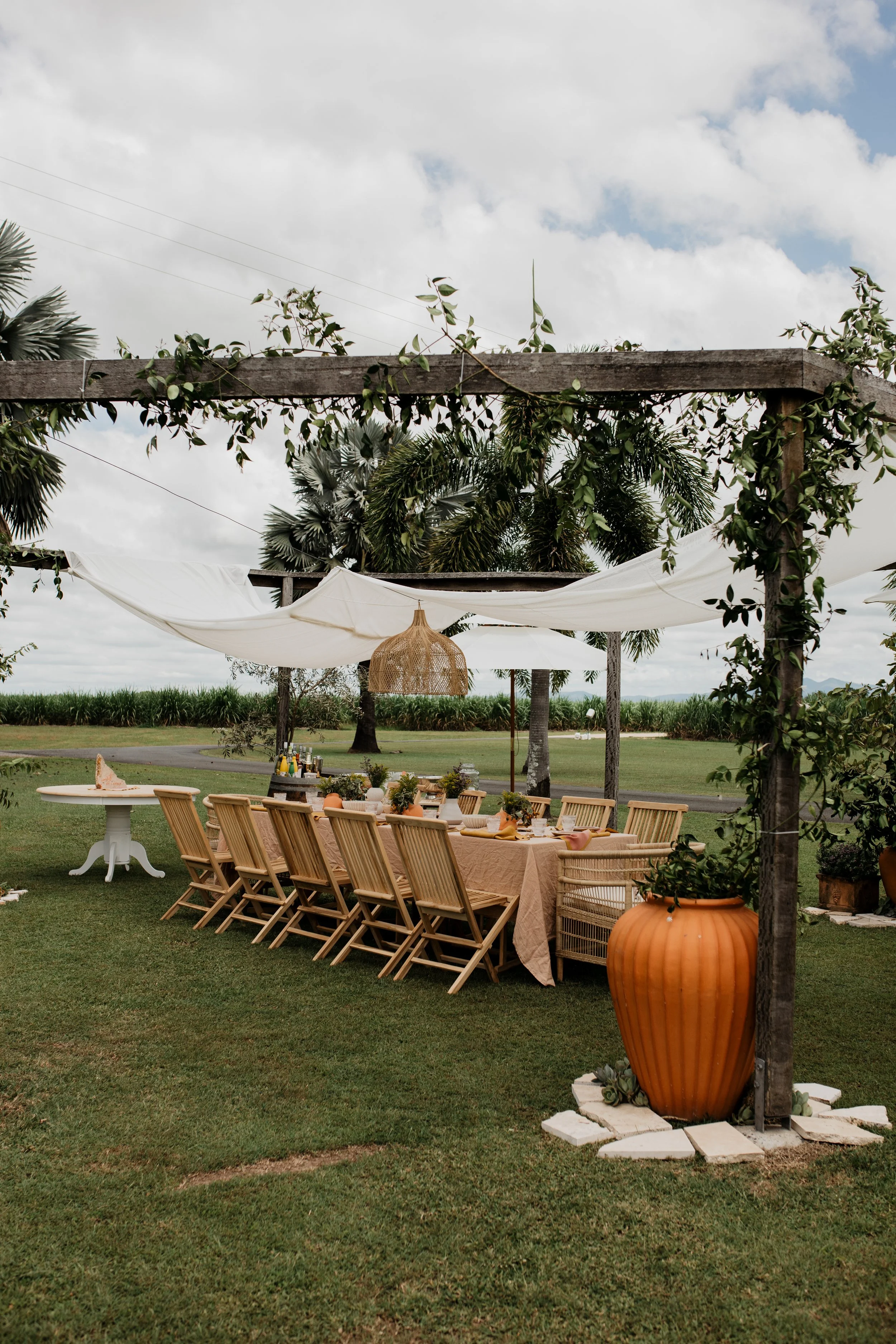 An outdoor dining setup with a long table covered with a beige tablecloth, surrounded by wooden chairs, under a pergola decorated with greenery and white fabric, with a large orange planter and lush green landscape in the background.