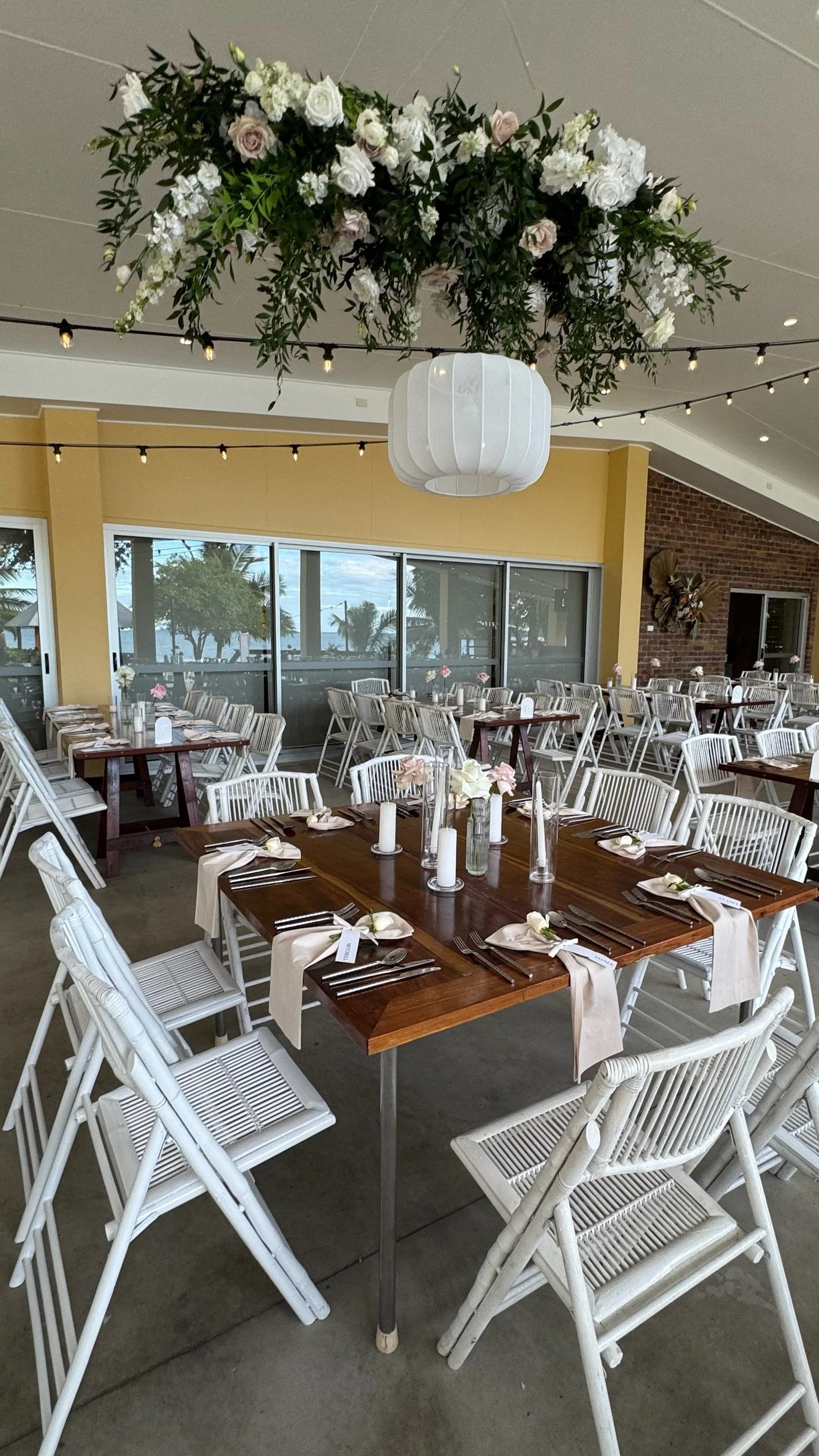 Elegant indoor event space with wooden tables, white chairs, floral centerpieces, candles, and a floral ceiling arrangement, overlooking a view of palm trees.