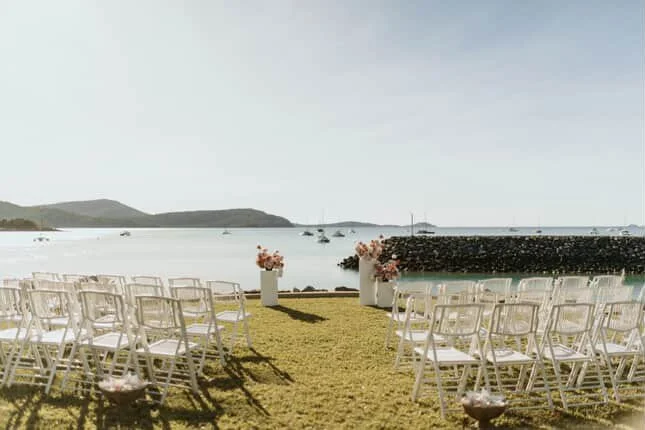 Outdoor wedding setup by the water with rows of white chairs, floral arrangements, and a backdrop of the sea and boats