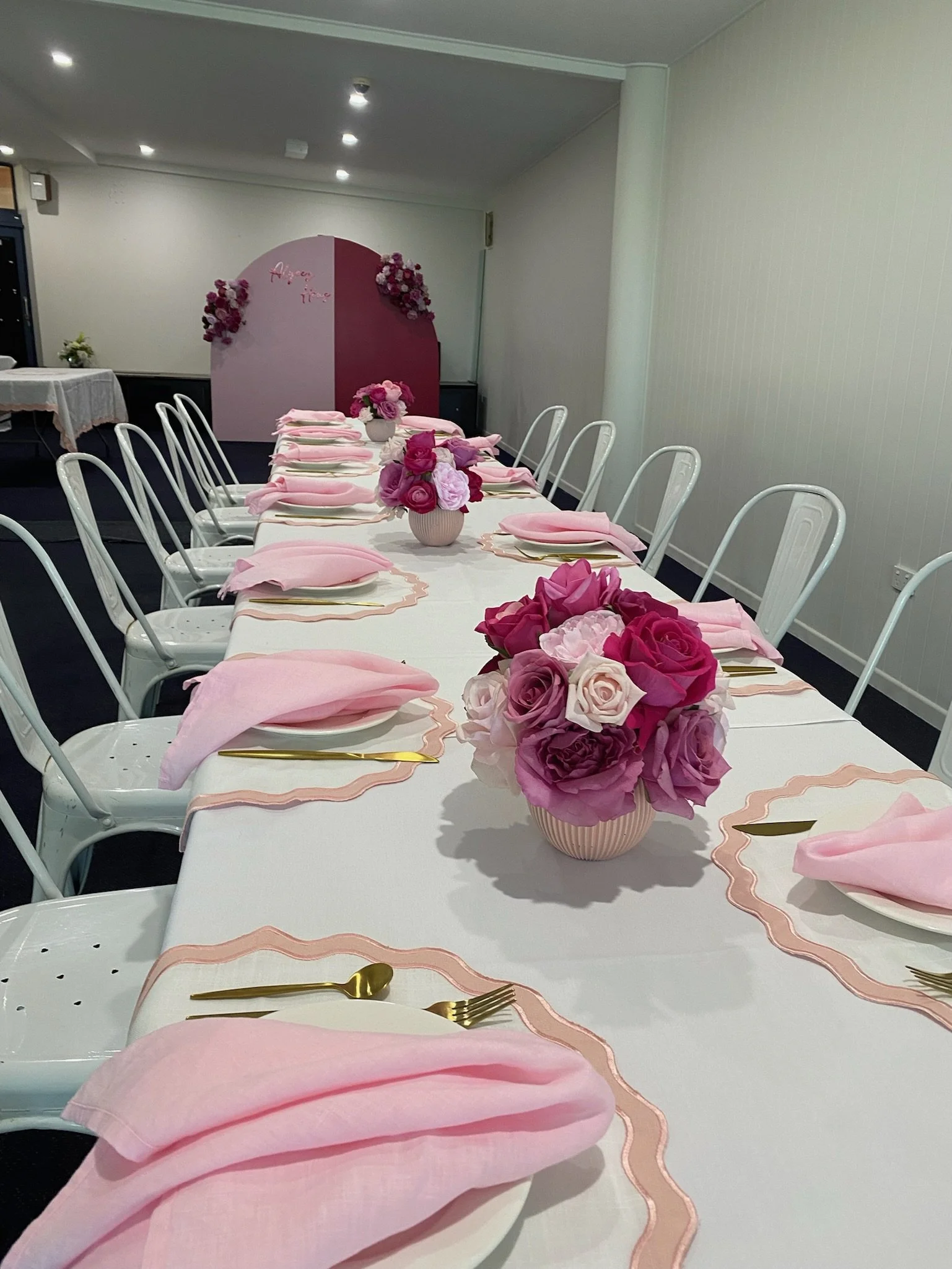 Decorated banquet table for a celebration with pink roses in vases, pink napkins, gold utensils, and a pink and white backdrop with floral decorations.
