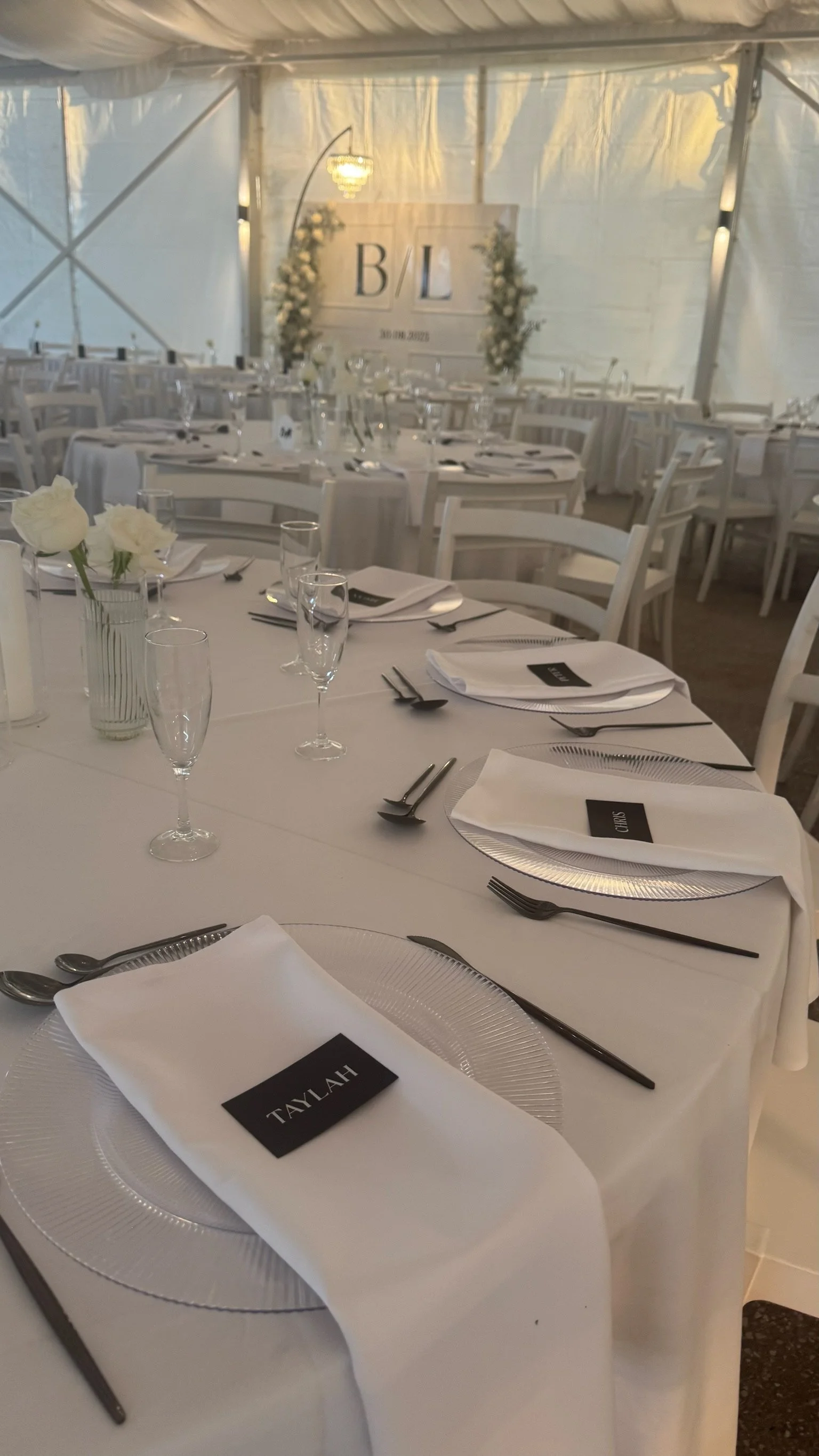 A decorated wedding reception table with white tablecloths, place settings, black name cards, glasses, and floral centerpieces, inside a tent with a sign reading 'B/L' and the date '30.08.2023' in the background.