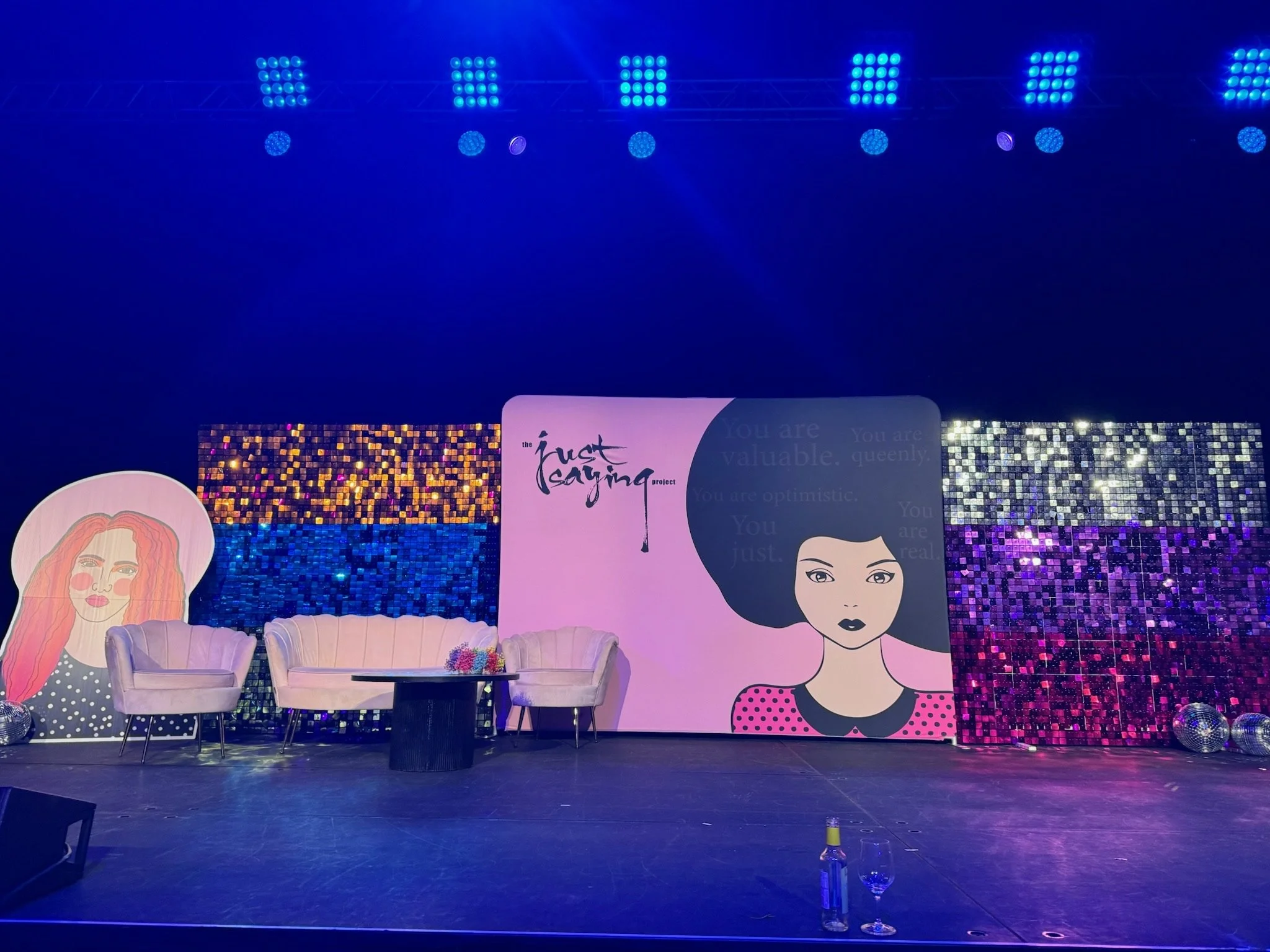 Stage setup with pink and purple decorations, a portrait of a woman with red hair, a large illustrated face of a woman with black hair, and a pink panel with text reading 'the trust saying project.' There are two white chairs, a round table with flow