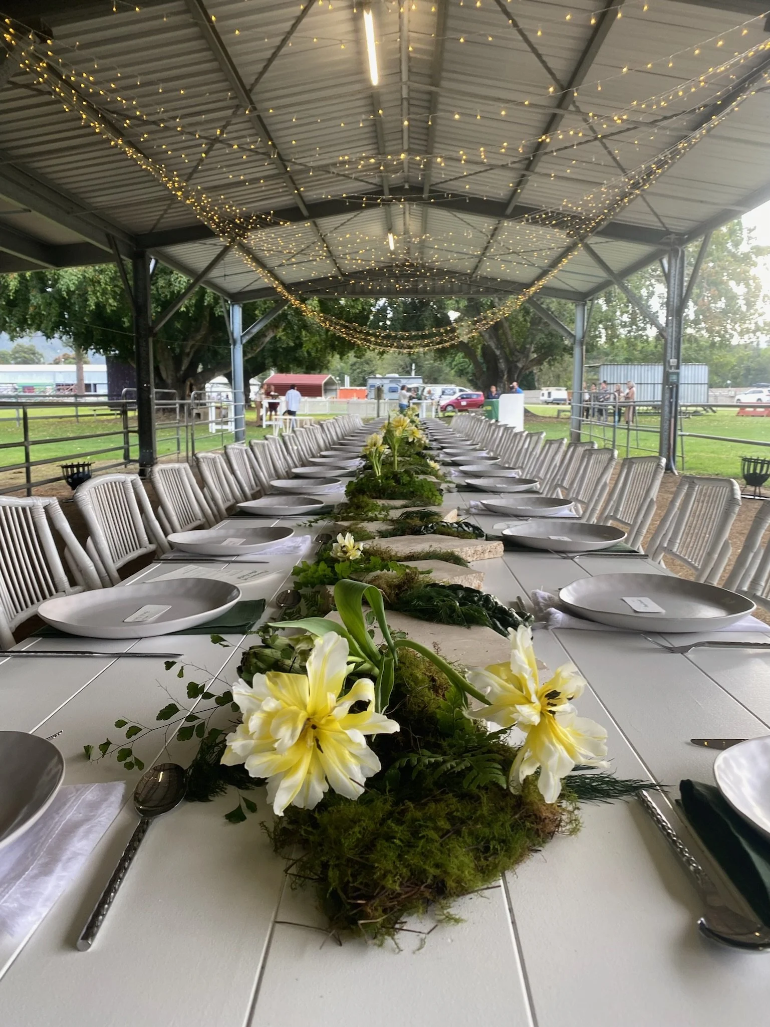 Long banquet table decorated with a lush green and white floral centerpiece, surrounding plates, and utensils. The table is under a metal pavilion with string lights overhead, set outdoors on a grassy area with trees and people in the background.