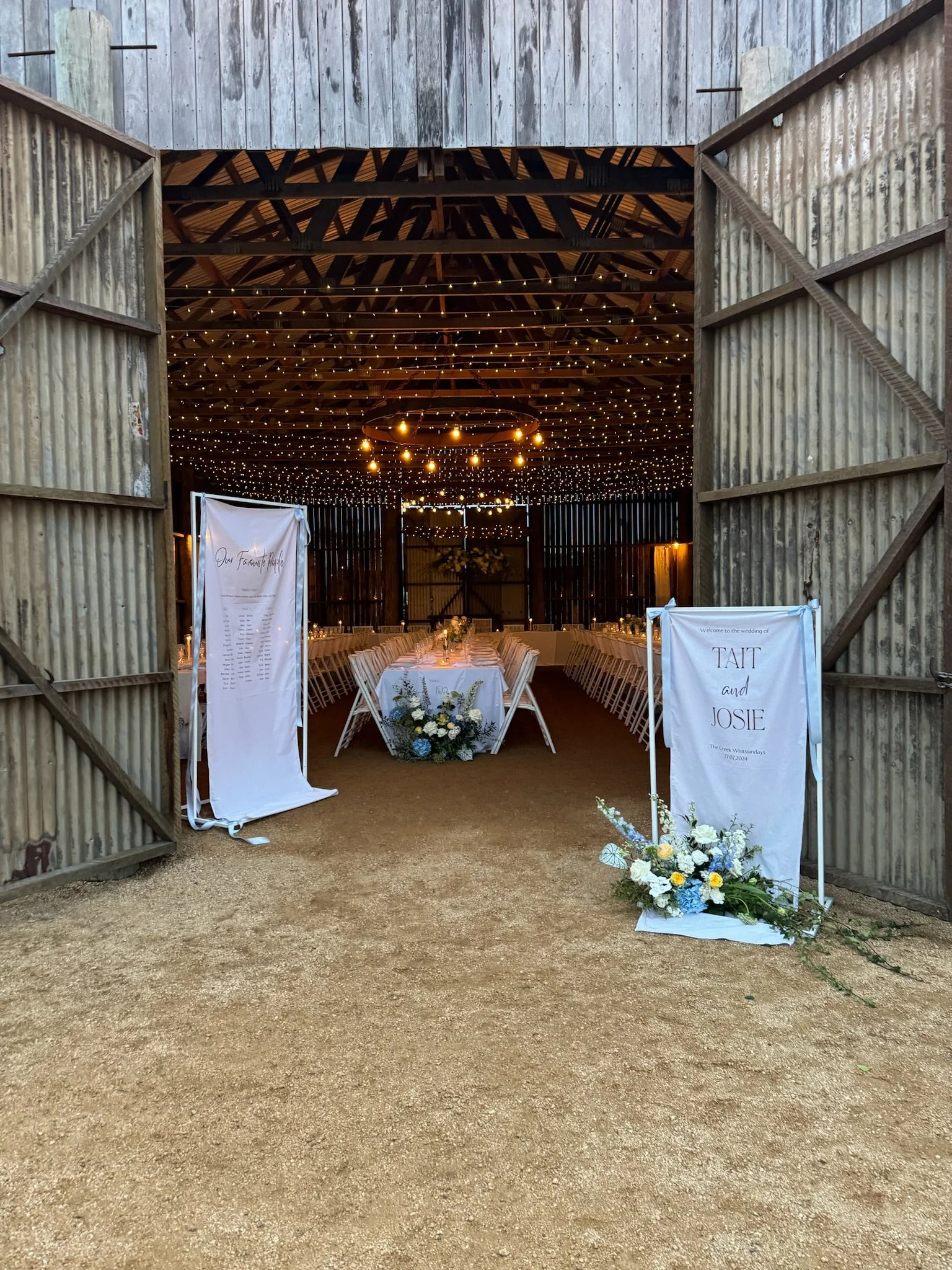 Open barn doors revealing decorated wedding reception area with string lights, long tables, floral arrangements, and signs with names 'Tait and Josie'.