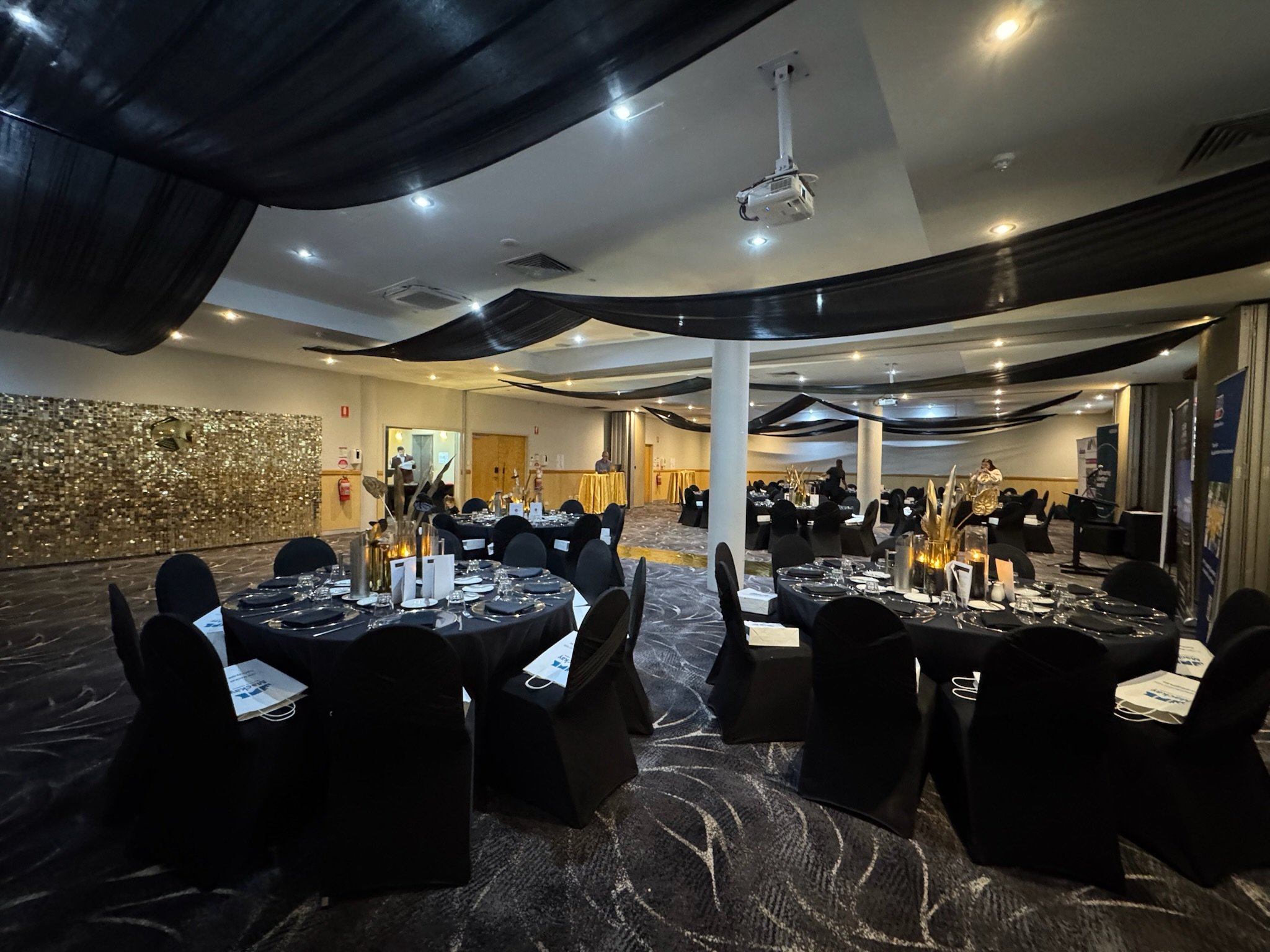 Event hall with round tables covered in black tablecloths, decorated with centerpieces, set for dinner, and black chairs. Black fabric drapes hang from the ceiling, and there are spotlights and a projector.