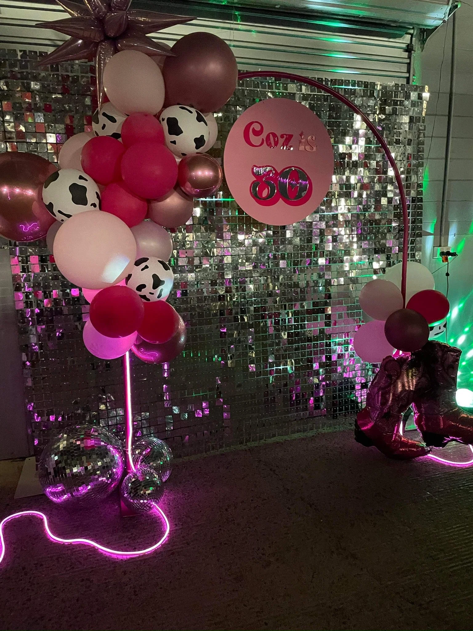 Decorative balloon arrangement celebrating Coz's 30th birthday, featuring pink, white, burgundy, and cow-print balloons, a pink circle sign with 'Coz is 30' in pink and silver, and a reflective mosaic backdrop with pink and green lighting.