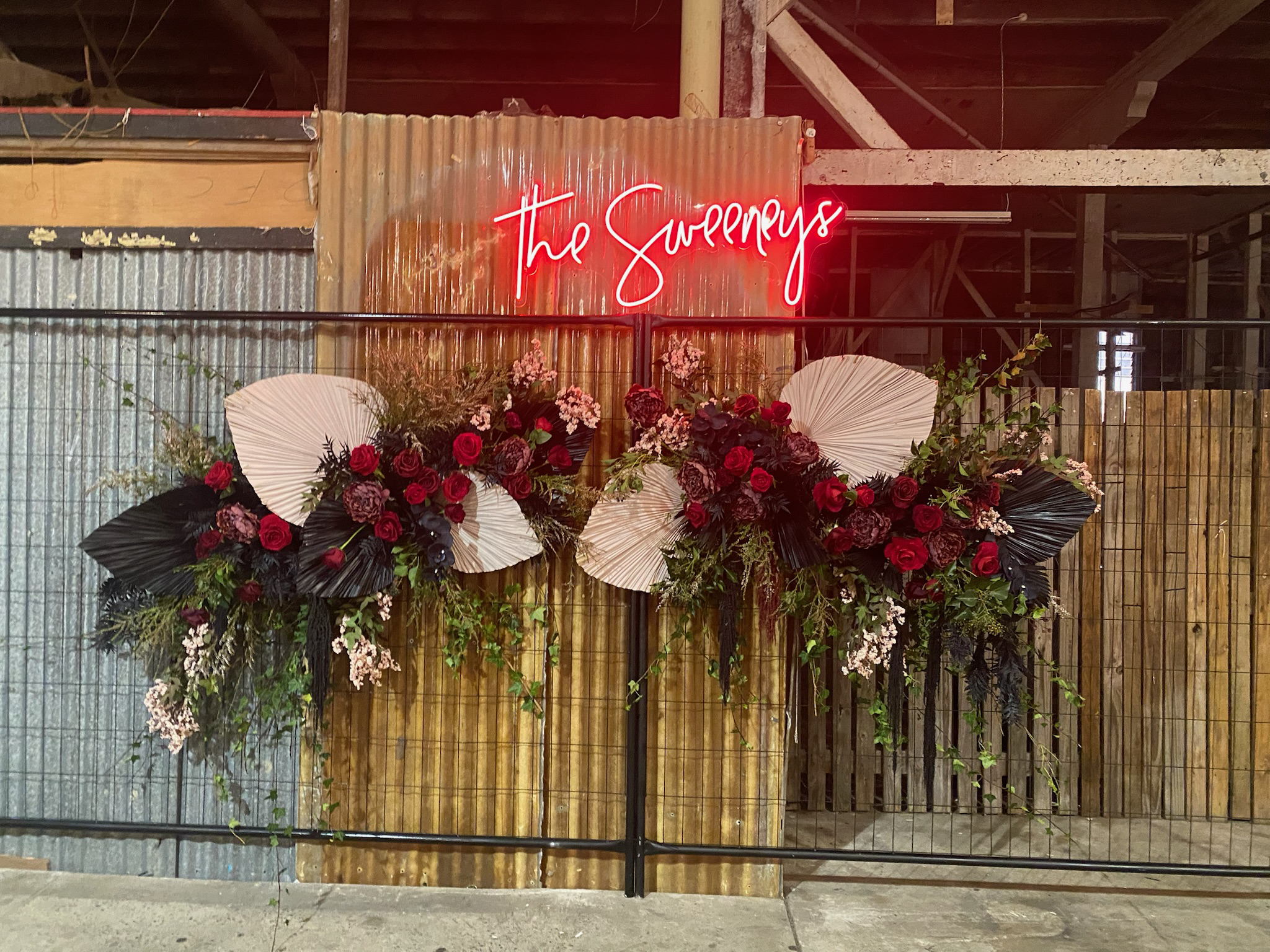 A floral display with red and dark purple flowers, white and black paper fan decorations, and greenery, beneath a red neon sign that reads 'The Sweeneys' in a rustic setting with wood and metal fencing.