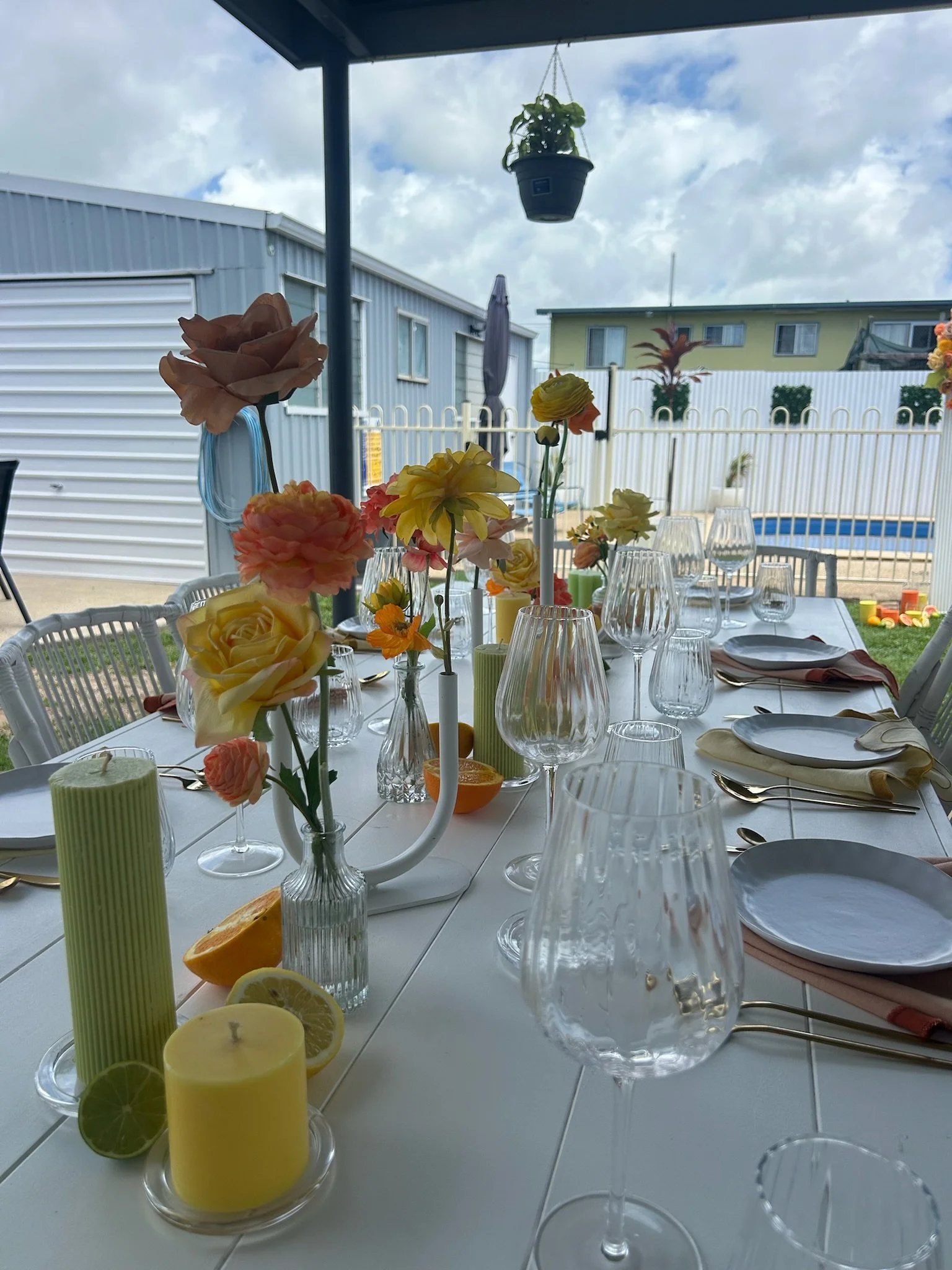 A decorated outdoor dining table with colorful flowers, candles, and glassware, set under a patio with a backyard and cloudy sky in the background.