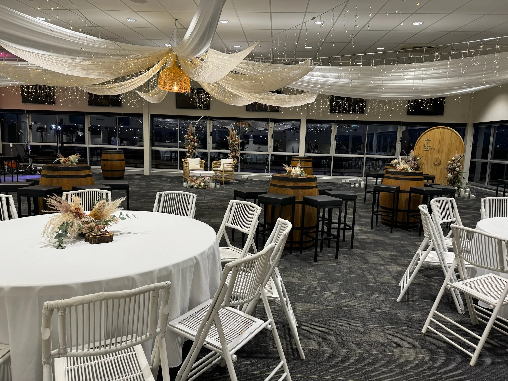 Event space decorated with white drapes and string lights on the ceiling. There is a round table with a floral centerpiece in the foreground, and the space includes chairs, barrels, and floral arrangements with a rustic and elegant theme.