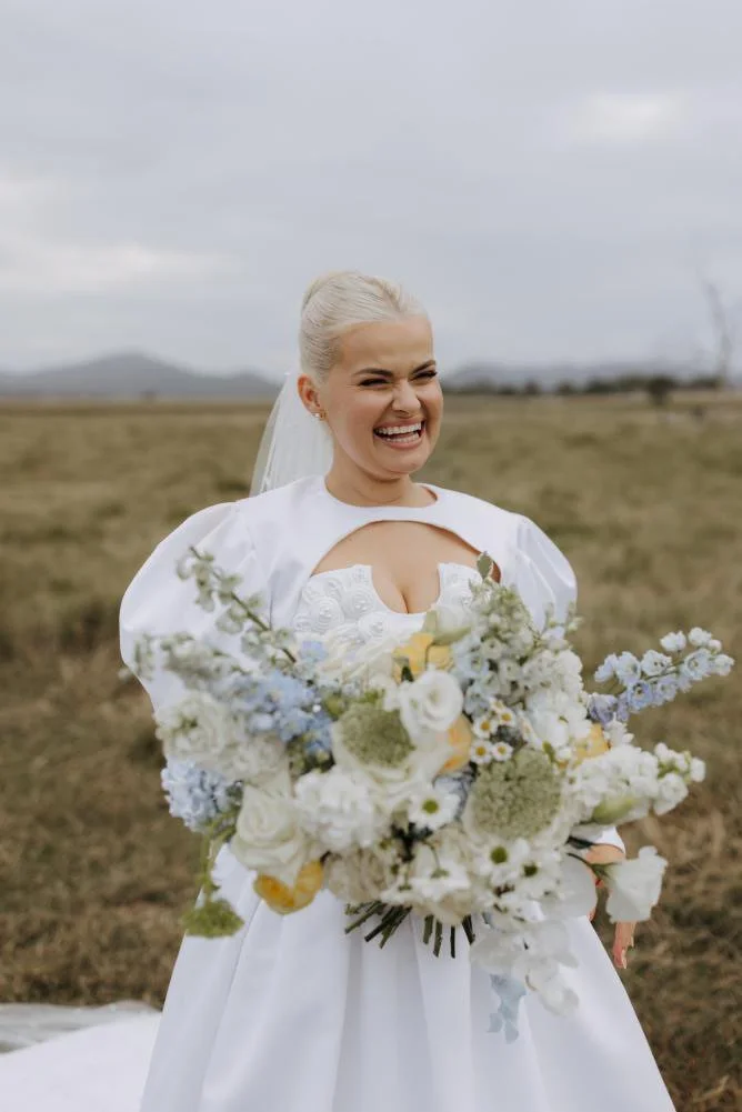A bride in a white wedding dress holding a large bouquet of white and light-colored flowers, smiling with a playful expression outdoors in a grassy field with a cloudy sky.