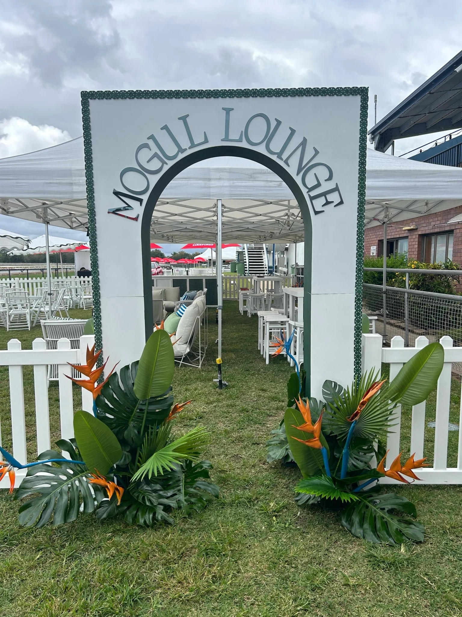 Entrance archway with 'Mogul Lounge' written on top, decorated with tropical plants and flowers, leading to outdoor seating area with white furniture and white fencing, under a cloudy sky.