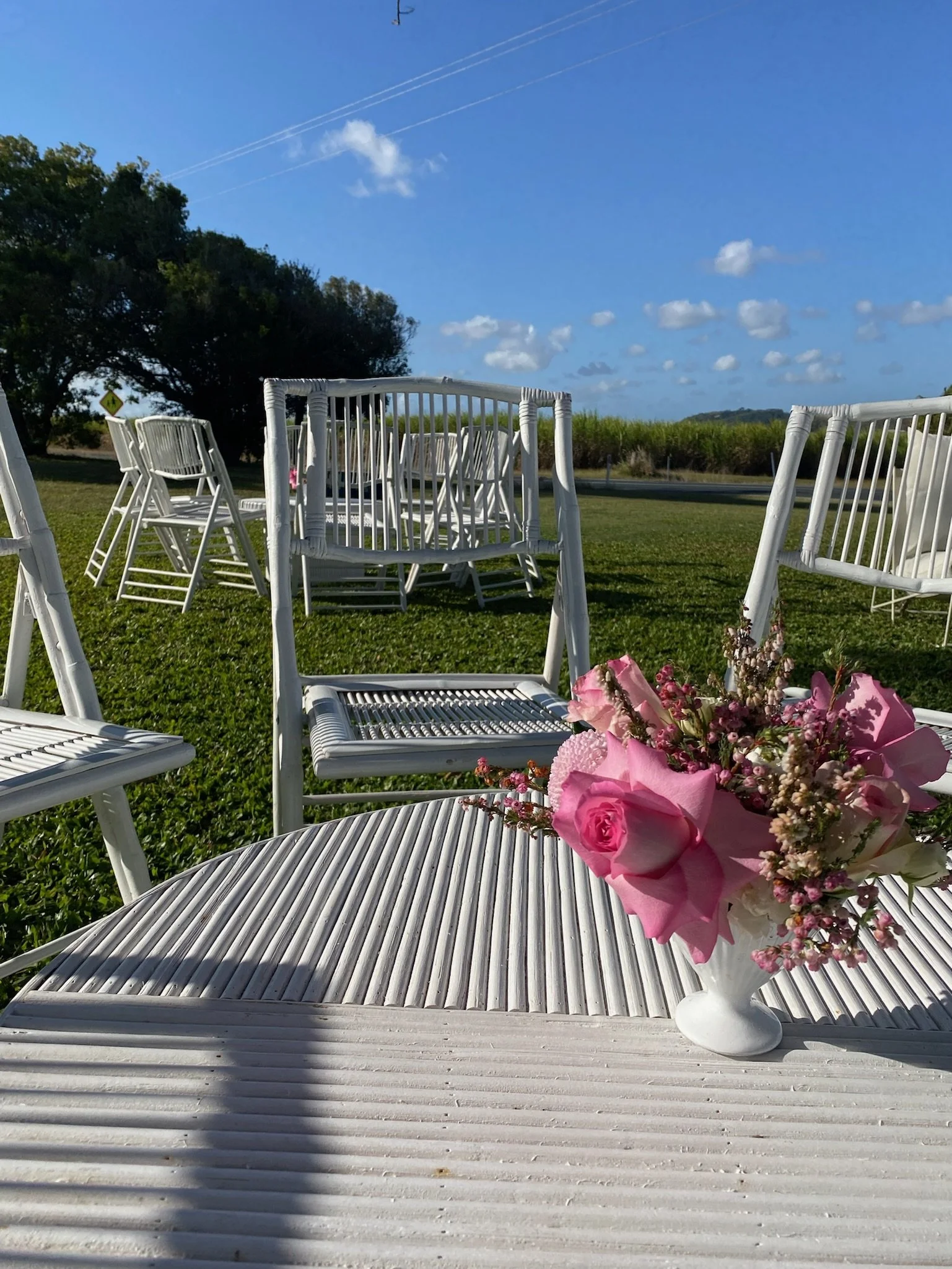 Outdoor scene with several white chairs arranged on green grass, a white table with pink and white flowers in a vase in the foreground, a blue sky with scattered clouds and contrails.