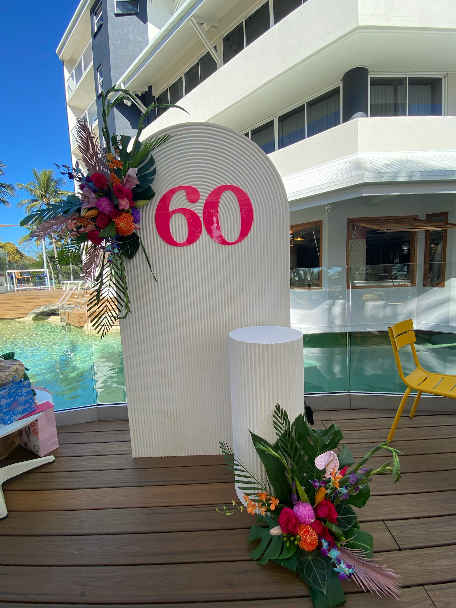 Decorative sign with the number 60 in pink, surrounded by colorful floral arrangements, set on a wooden deck near a swimming pool with a modern building in the background.