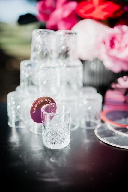 A set of clear glass drinking glasses, stacked in a pyramid, with one glass in front labeled "Ladies Night" and decorated with pink flowers in the background.