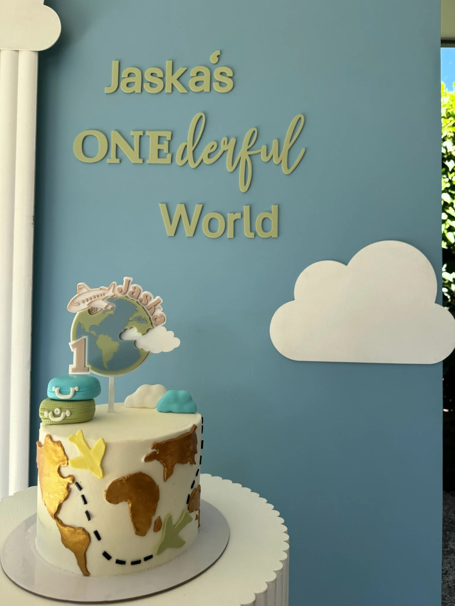 A themed cake decorated with a world map, an airplane, clouds, and travel-related decorations, in front of a blue background with the text 'Jaska's ONEderful World' and clouds.