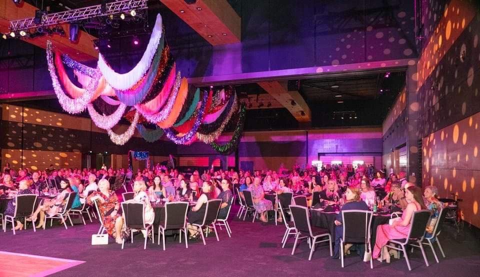 Large indoor event with many attendees seated at round tables, decorated with colorful hanging streamers and purple lighting, in a modern venue.