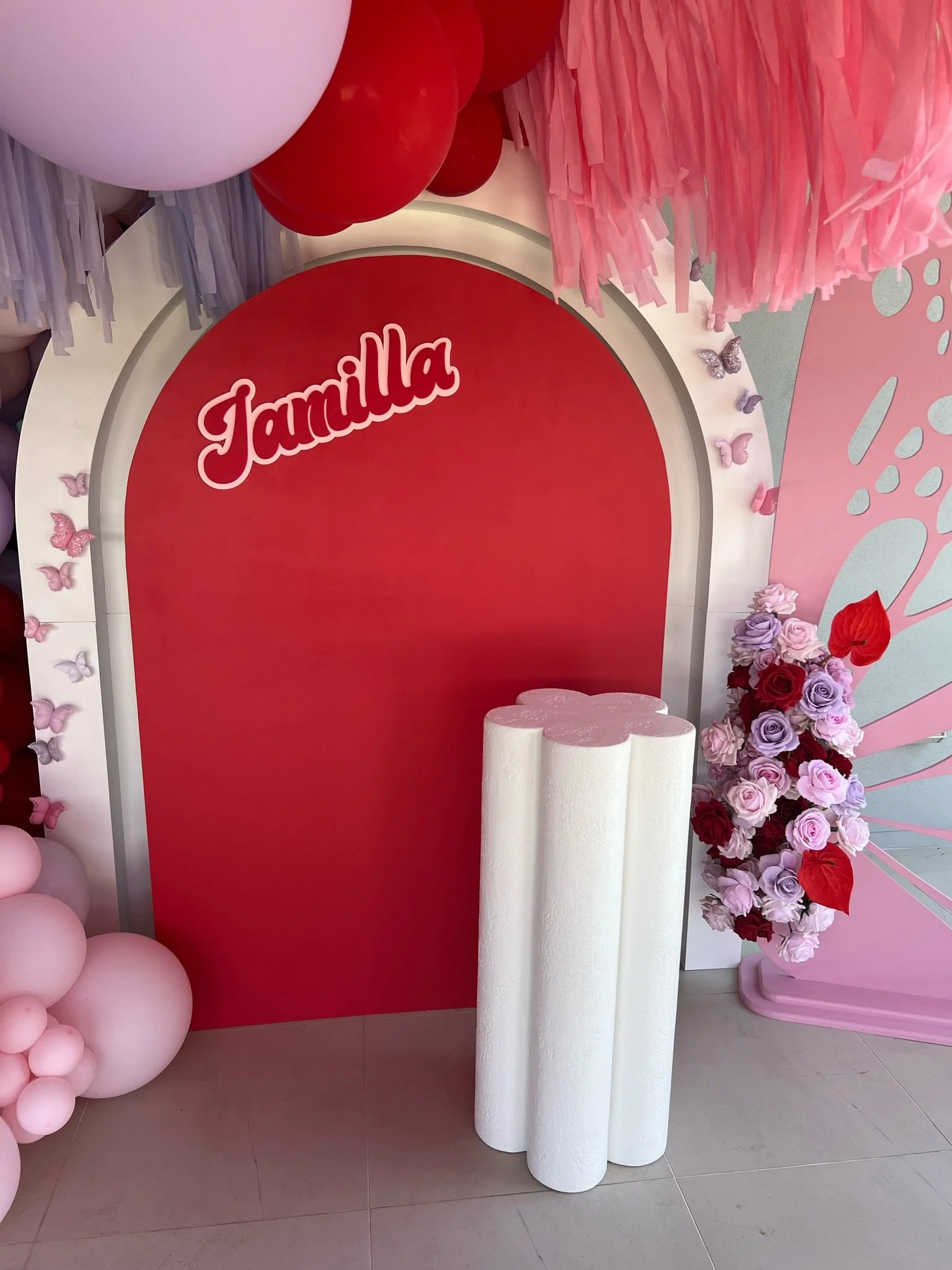 Decorative backdrop with the word 'Jamilya' in red cursive font, pink, red, and purple floral arrangements, pink balloons, and white cylindrical columns for a celebration or event.