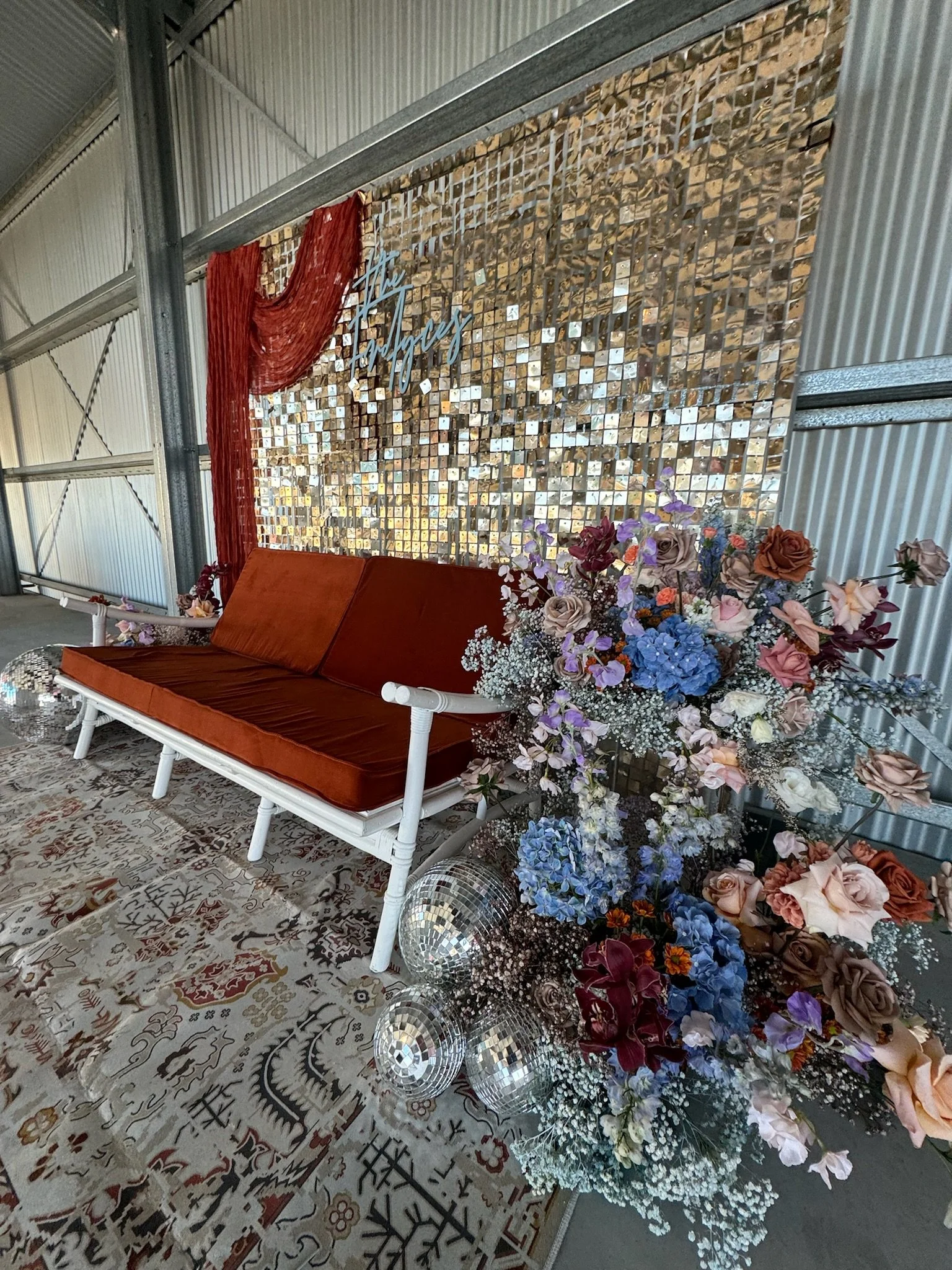 A decorative seating area with a white framed sofa with burnt orange cushions, a large floral arrangement with pink, purple, blue, and cream flowers, a mirrored wall with the words 'The Grooms' in cursive, disco balls on the floor, and patterned rugs