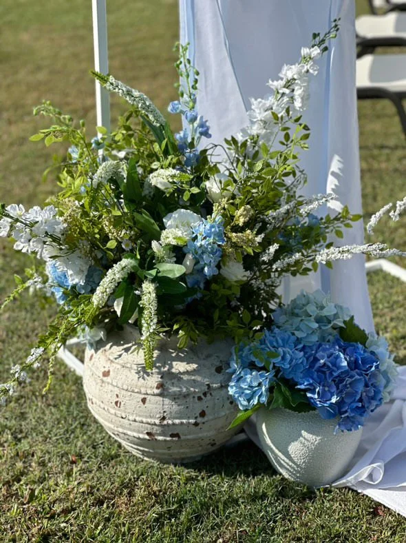 A floral arrangement with white and blue flowers in a large white vase outdoors, with green grass and a white fabric in the background.