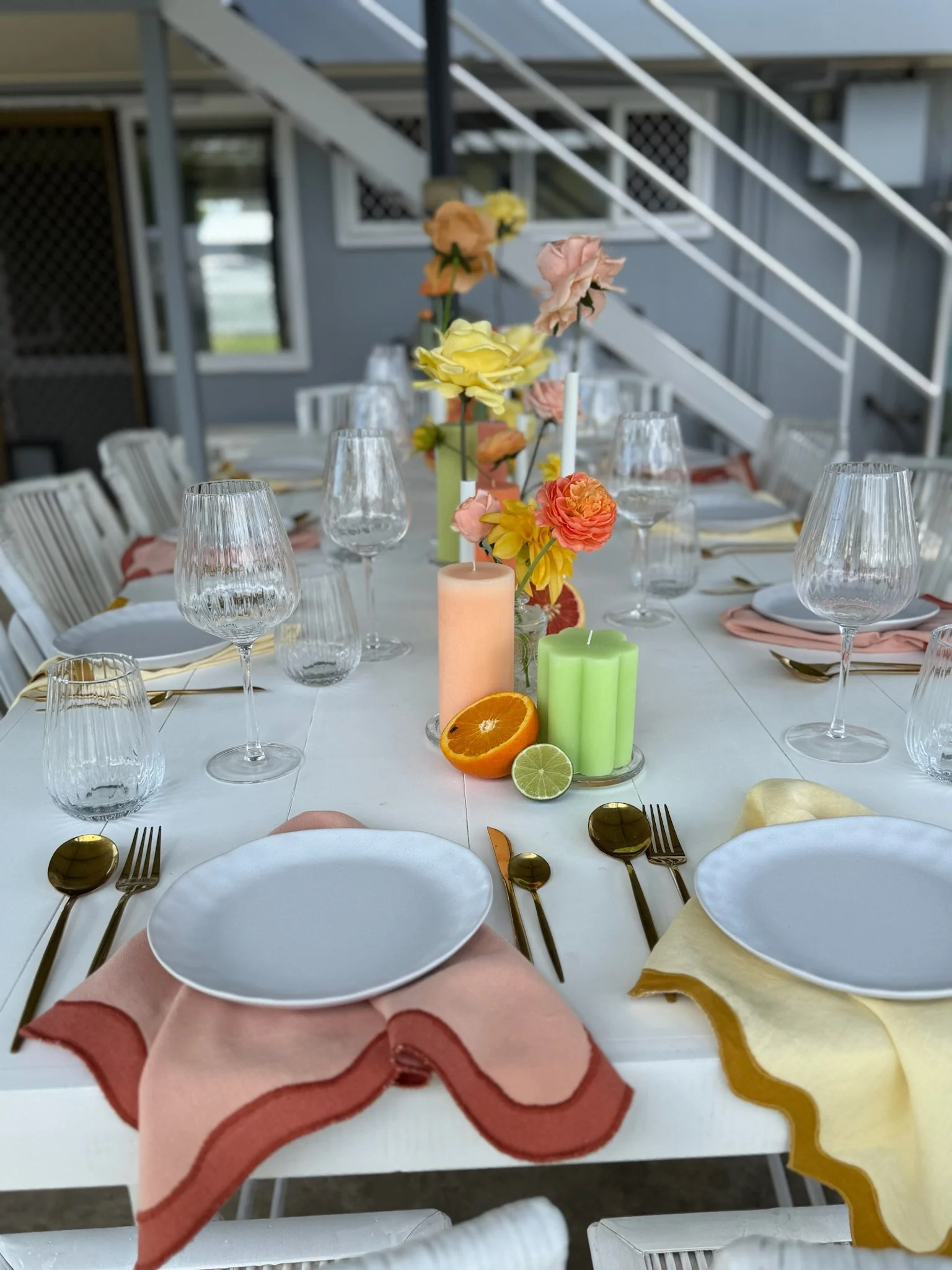 Decorated outdoor dining table with pastel-colored napkins, glasses, plates, and a floral centerpiece with candles and sliced citrus fruits.